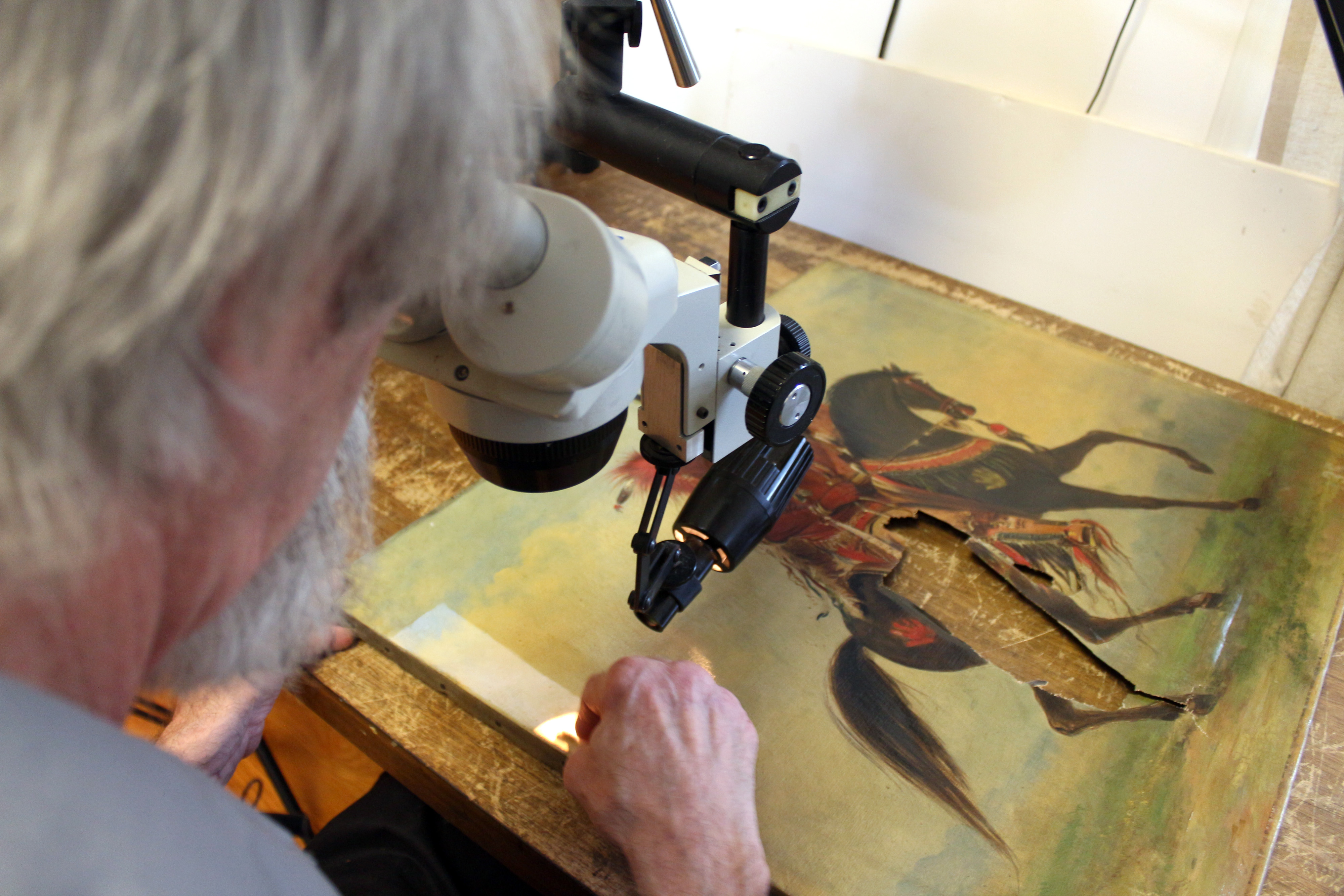 In this Wednesday, Aug. 26, 2015 photo, in River Forest, Ill., painting conservator Barry Bauman works in his studio cleaning a George Catlin painting owned by the State Historical Society of Iowa while looking through a binocular microscope. He's cleaned and fixed 1,500 paintings for hundreds of museums nationwide over the 11 years and he's done it all for free. (AP Photo/Carrie Antlfinger)