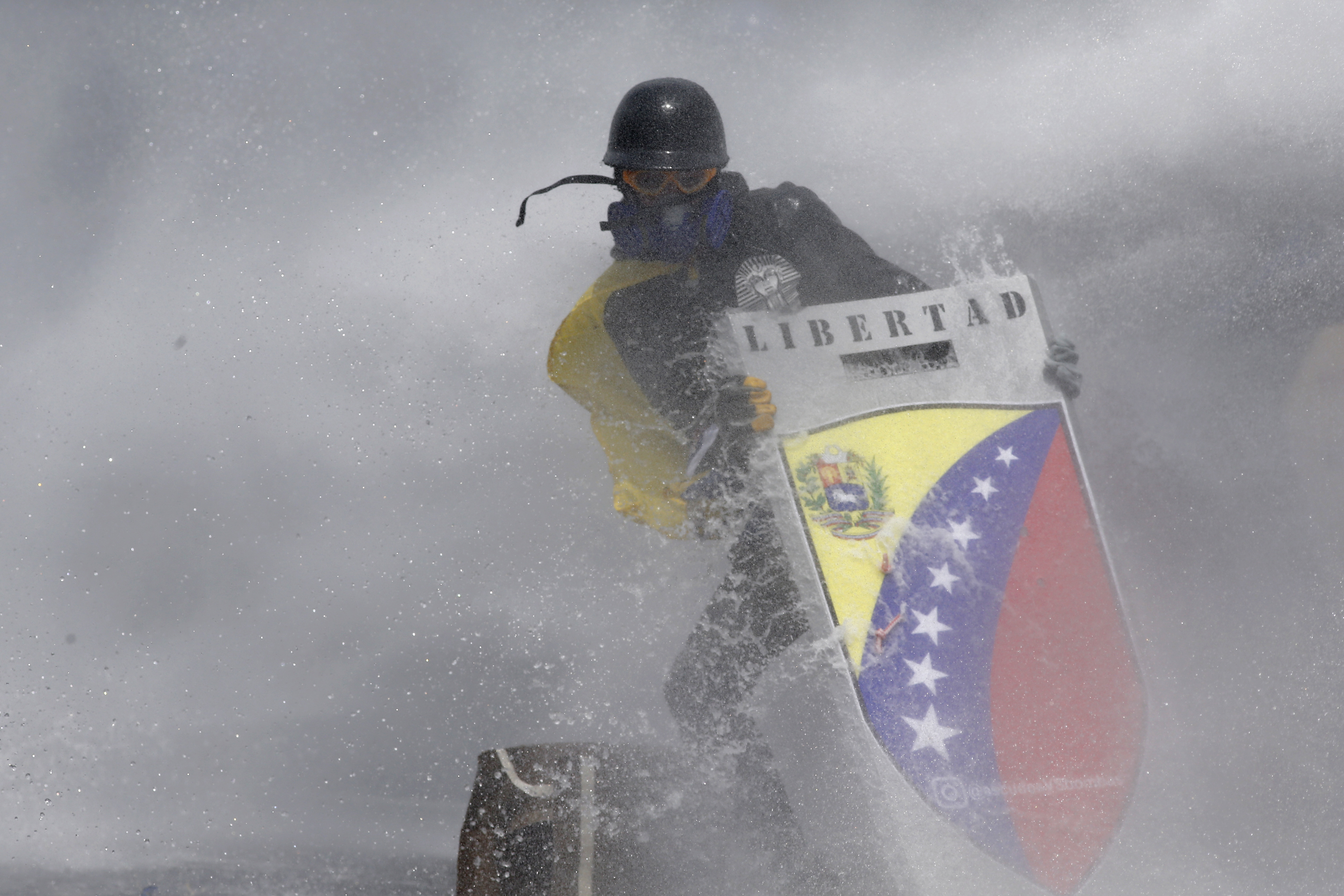 FILE - In this May 10, 2017 file photo, amid a cloud of tear gas, an anti-government protester wearing a helmet, googles, gas mask and gloves holds a shield with the Spanish word "Freedom," to protect himself from a water cannon fired by security forces in Caracas, Venezuela. The government's response to the demonstrations has drawn international condemnation, with U.S. Secretary of State Rex Tillerson expressing concern in April that Maduro is "not allowing the opposition to have their voices heard." (AP Photo/Ariana Cubillos, File)