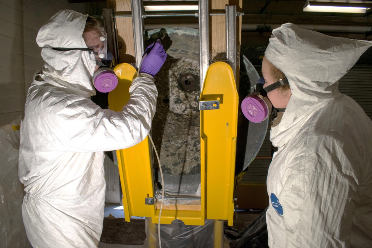 Melissa Swanson and Karl Knauer, conservators at Naval History and Heritage Command, conduct micro-abrasive cleaning treatments on the underside of the USS Utah bell's rim. (U.S. Navy photo by Naval History and Heritage Command Collection Management Facility staff)
