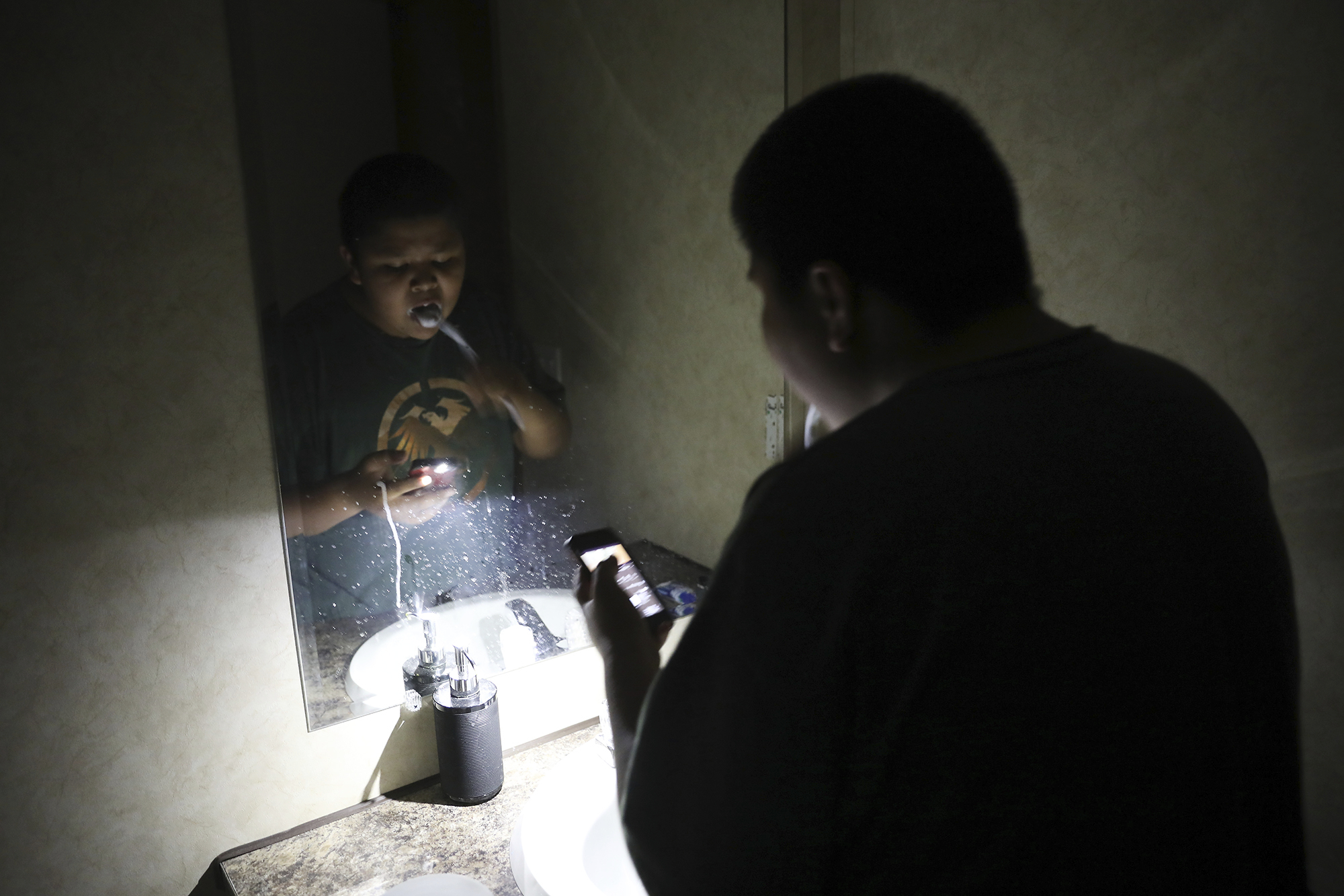 In this Wednesday, May 8, 2019 photo, Jayden Long, 13, brushes his teeth by cell phone light in the bathroom of his home on the Navajo Reservation. Long has spent more than a decade living with his family in a home with no electricity relying on a generator for power. (AP Photo/Jake Bacon)