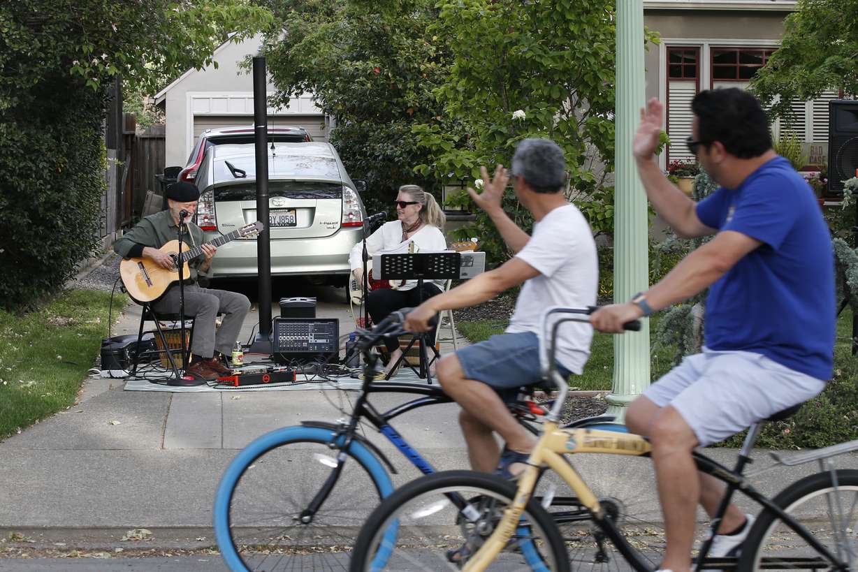 A pair of bicyclists, foreground, wave to musicians George Sheldon, left, and Sandra Carter as they perform a neighborhood concert in Sacramento, Calif., Friday, April 24, 2020. With people following the mandatory stay-at- home directive, the duet was invited by friends to perform a Friday night show. The pair that performs locally by the name "Be Here Now," has had several appearances canceled due to the coronavirus pandemic. (AP Photo/Rich Pedroncelli)