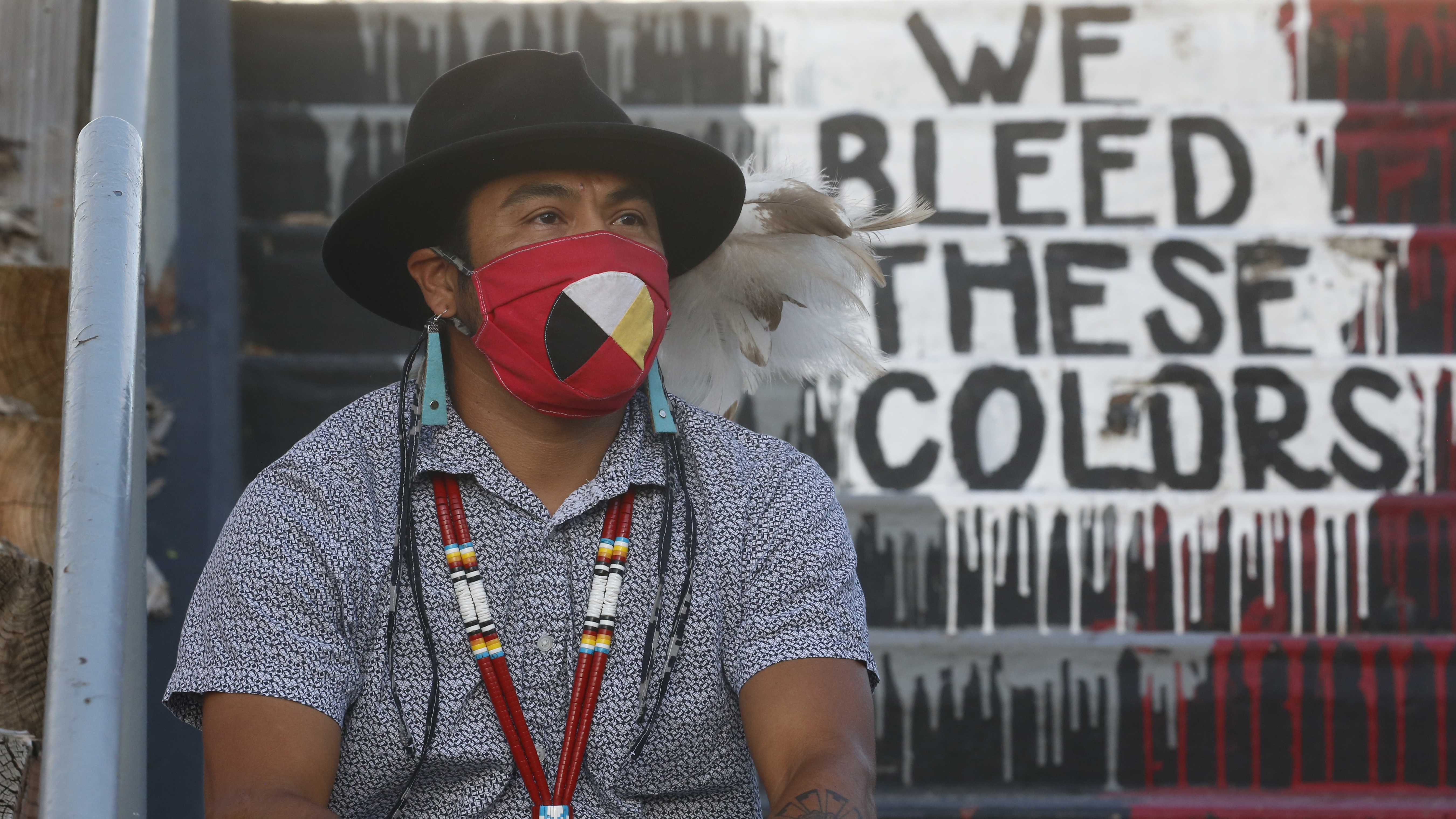 Native American advocate Carl Moore sits near the phrase "We Bleed These Colors" along a walkway which leads from the Bountiful High School parking lot up to the football field Tuesday, July 28, 2020, in Bountiful, Utah. While advocates have made strides in getting Native American symbols and names changed in sports, they say there's still work to do mainly at the high school level, where mascots like Braves, Indians, Warriors, Chiefs and Redskins persist.
