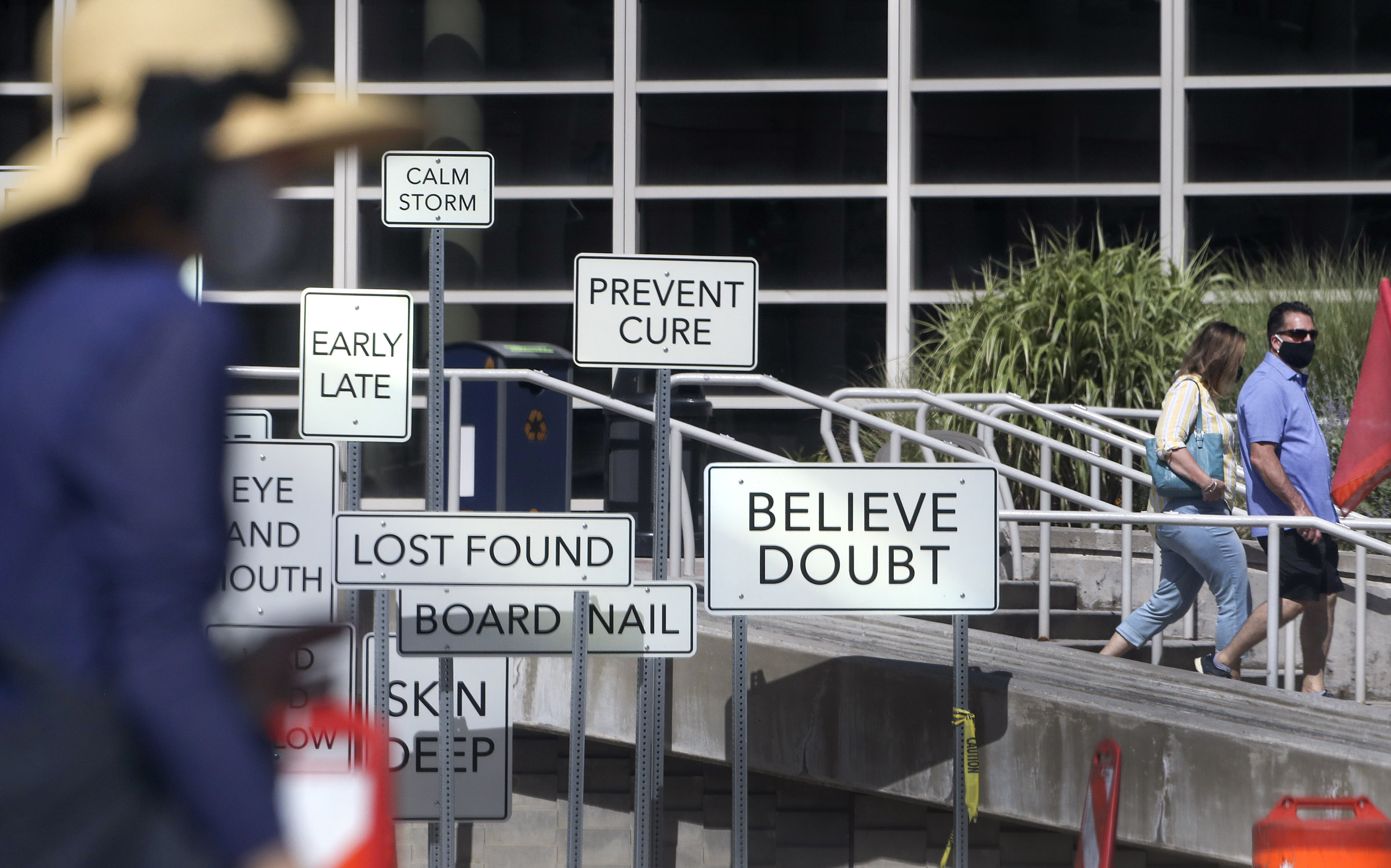 Pedestrians wear masks as they walk past the “Point of View” art installation in front of the Salt Palace Convention Center in Salt Lake City on Monday, July 27, 2020. The installation, at West Temple and 100 South, was created by Maine artist Aaron T Stephan and features over 150 road signs with diametrical words on them.