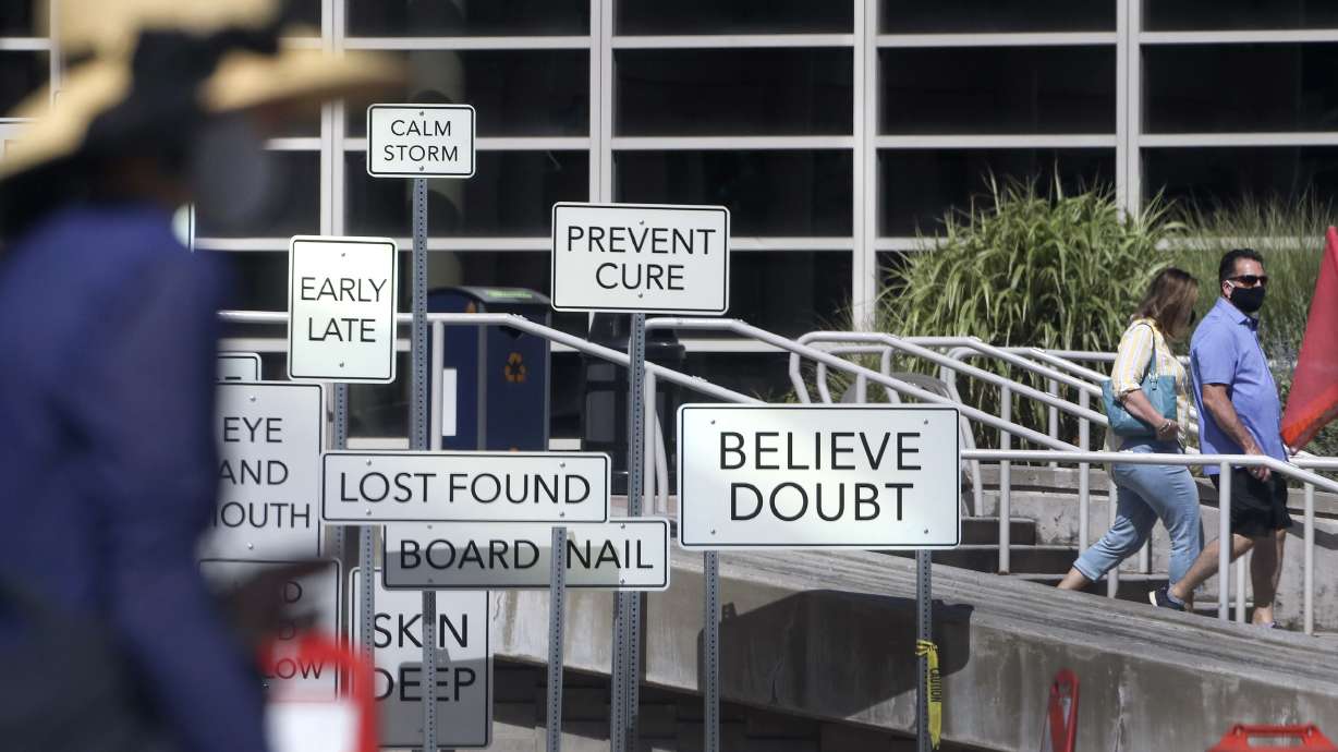 Pedestrians wear masks as they walk past the “Point of View” art installation in front of the Salt Palace Convention Center in Salt Lake City on Monday, July 27, 2020. The installation, at West Temple and 100 South, was created by Maine artist Aaron T Stephan and features over 150 road signs with diametrical words on them.