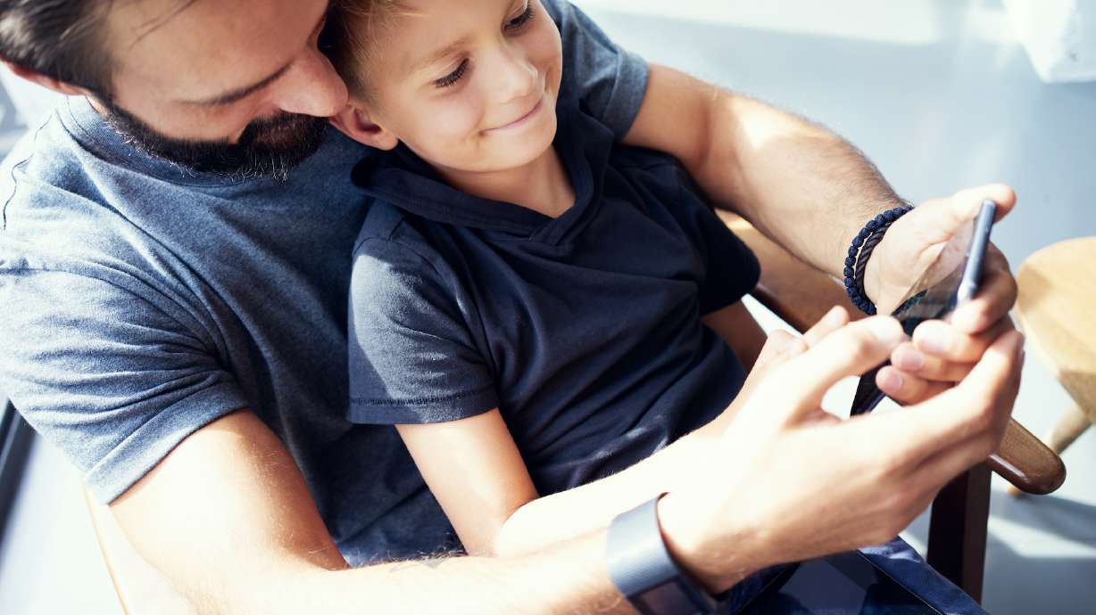 Closeup of young boy sitting with father and using mobile phone in modern sunny place. Horizontal, blurred background
