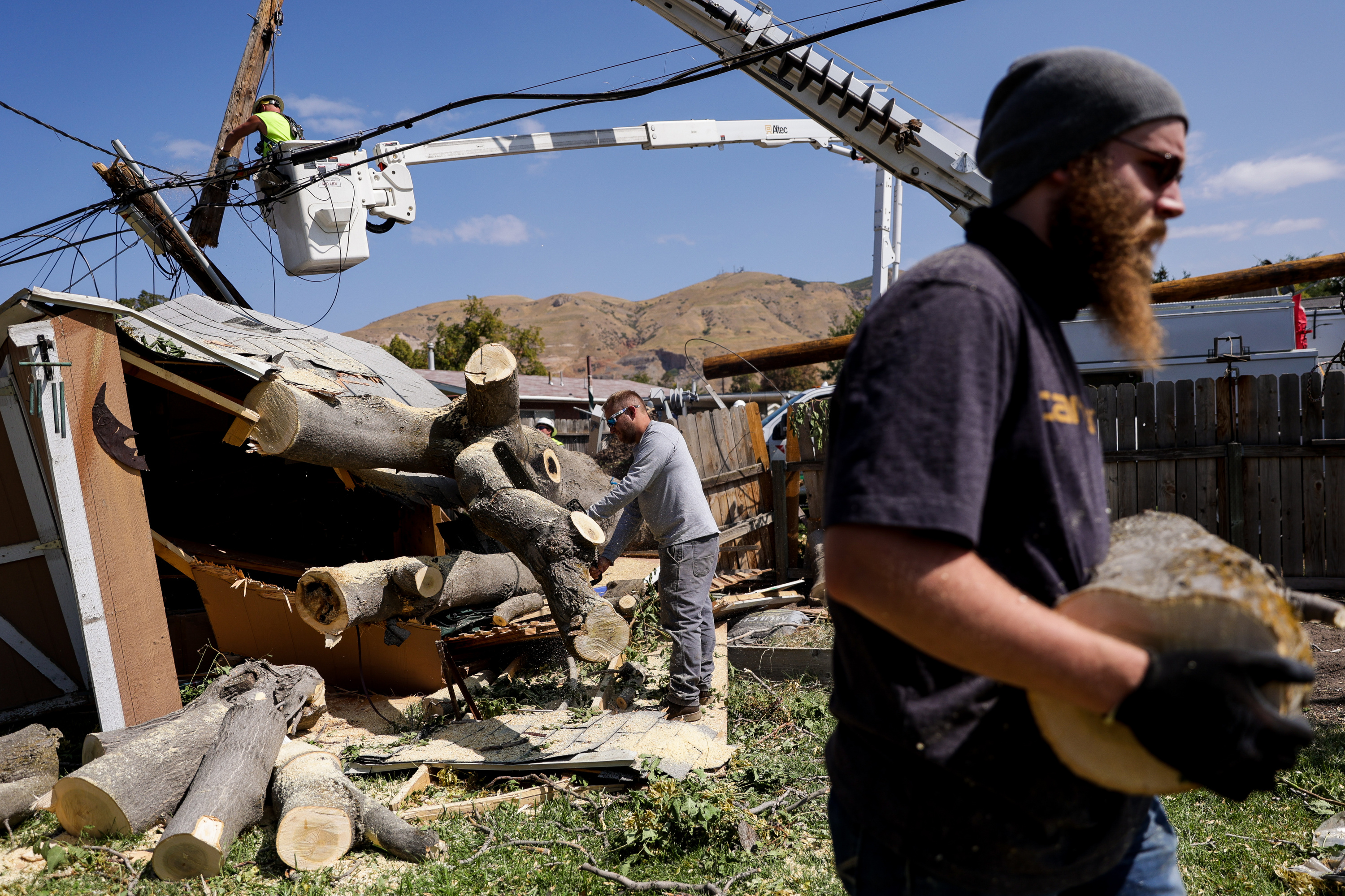 James Denton, center, and Conrad Kleinman, right, cut up a tree that smashed a shed in the yard of Dentonâ??s mother, Laura Lee, in the Rose Park neighborhood of Salt Lake City on Thursday, Sept. 10, 2020. In the background, crews from MidAmerican Power, an Iowa-based power company, works to fix a downed power pole and lines. Residents and utility companies are continuing to clean up after severe winds hit the Wasatch Front on Tuesday and Wednesday, leaving hundreds of thousands without power.
