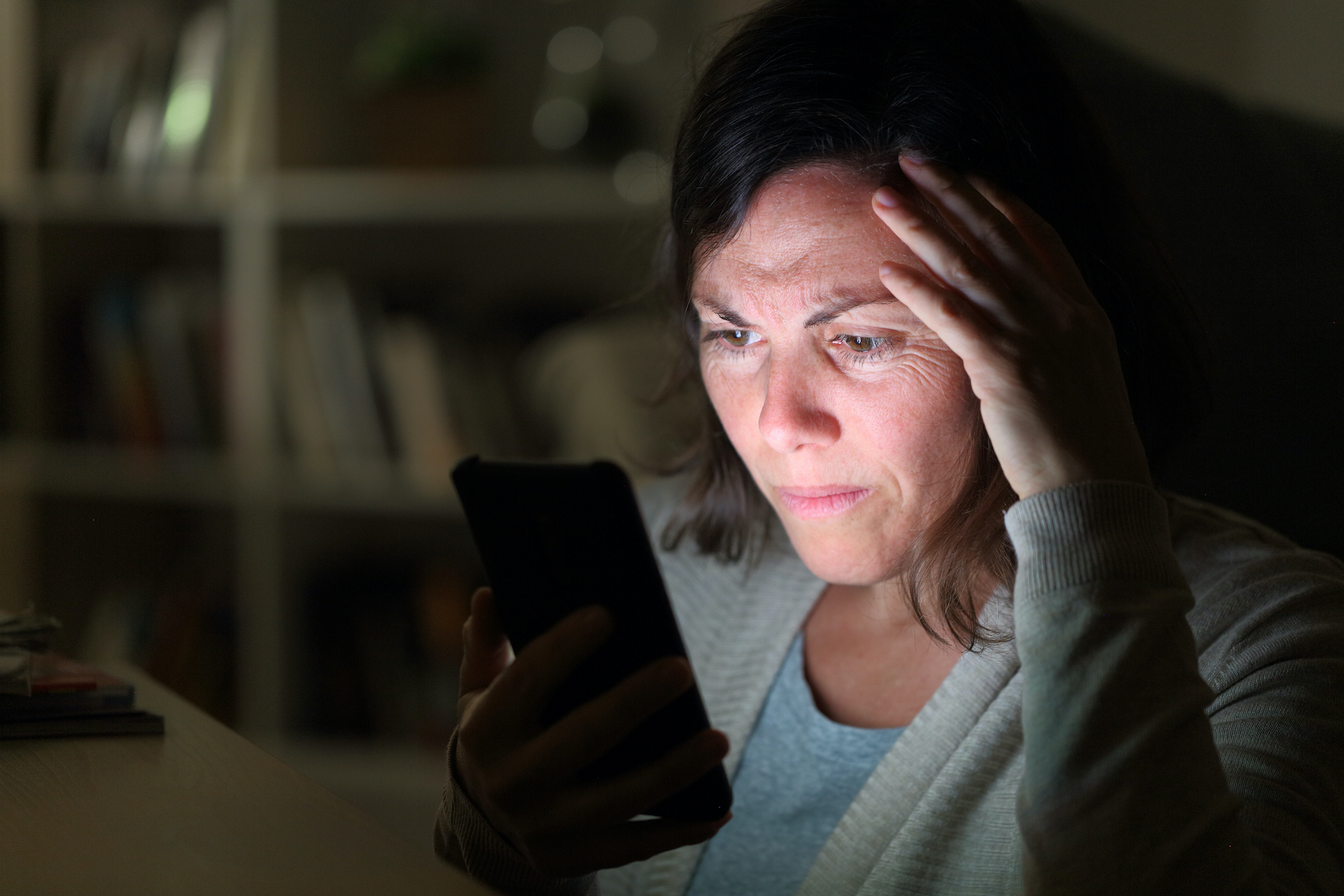 Preoccupied adult woman reading on smartphone lighted screen sitting at night at home