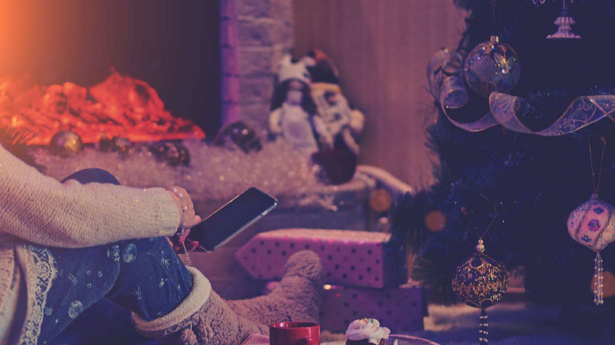 People, Christmas, holidays, communication, technology and internet concept. Woman with smartphone at home on the fireplace and Christmas tree background. Red mug of hot chocolate and gingerbread.