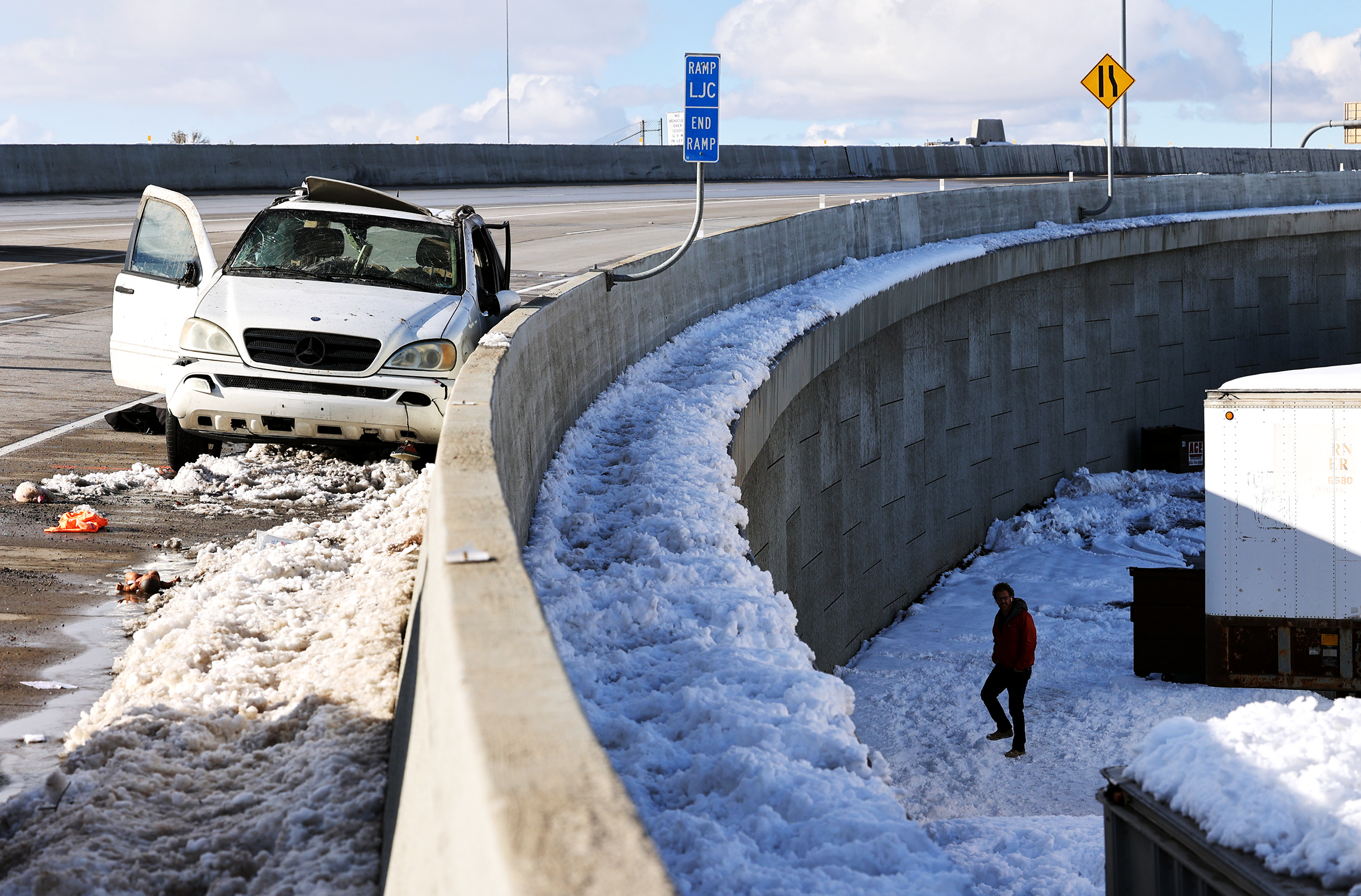 A white Mercedes SUV involved in a fatal rollover sits against a concrete barrier on the 500 South on-ramp to westbound I-80 as a man searches an area below in Salt Lake City on Wednesday, Feb. 17, 2021. According to officials, the SUV being driven by the woman was traveling northbound on I-15 at 500 South and the I-80 westbound on-ramp. The driver of the SUV merged from the far left lane on I-15 and attempted to exit onto the I-80 westbound connecting ramp. In doing so, the driver lost control and rolled at least twice. The SUV came to rest on the 500 South on-ramp to westbound I-80 westbound. The female driver was ejected from the SUVâs sunroof and came to rest after falling roughly 30 feet from the raised freeway structure. A male passenger and two children in the back seat were transported to the hospital as a precaution but appeared to be uninjured in the crash.