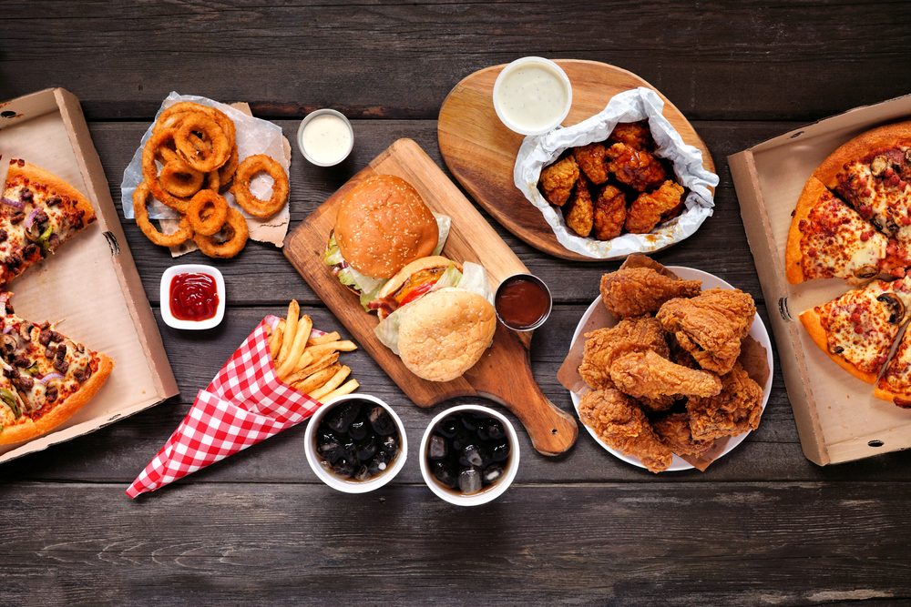 Table scene with large variety of take out and fast foods. Hamburgers, pizza, fried chicken and sides. Above view on a dark wood background.