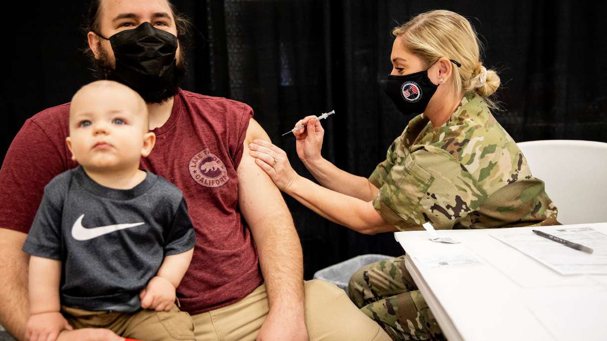 Isaac Hoffman, with his 10-month-old son Leo sitting on his lap, receives a dose of the Pfizer-BioNTech COVID-19 vaccine from Utah Air National Guard Master Sgt. Lauren Farley at a vaccination site run by the Utah County Health Department in Spanish Fork on Thursday, March 25, 2021.