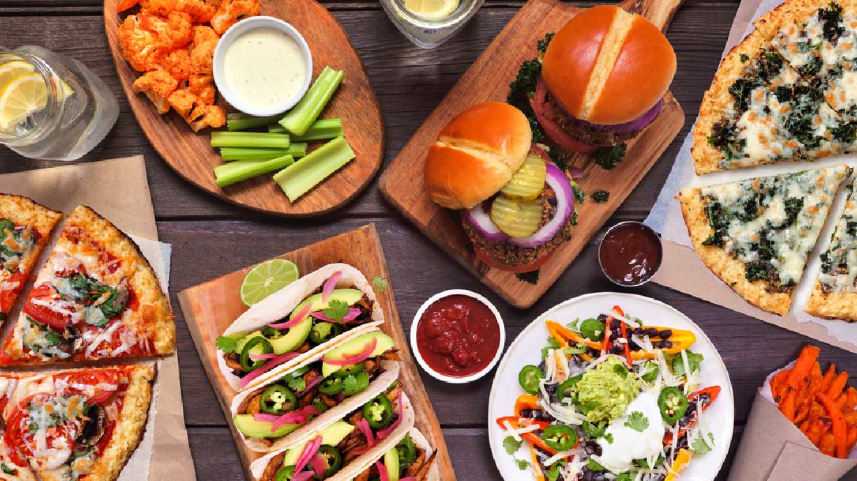 Healthy plant based fast food table scene. Overhead view on a wood background. Cauliflower crust pizzas, bean burgers, mushroom tacos, bell pepper nachos and cauliflower wings and sweet potato fries.