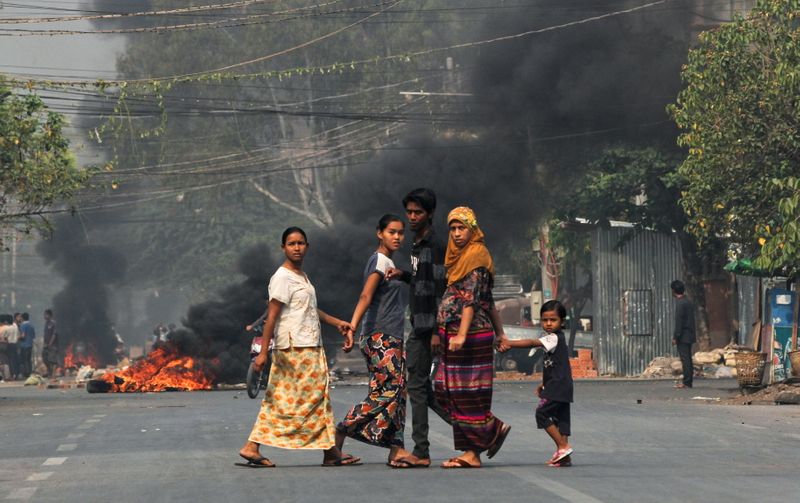 People walk on a street as barricades burn behind them during a protest against the military coup, in Mandalay, Myanmar March 27, 2021.