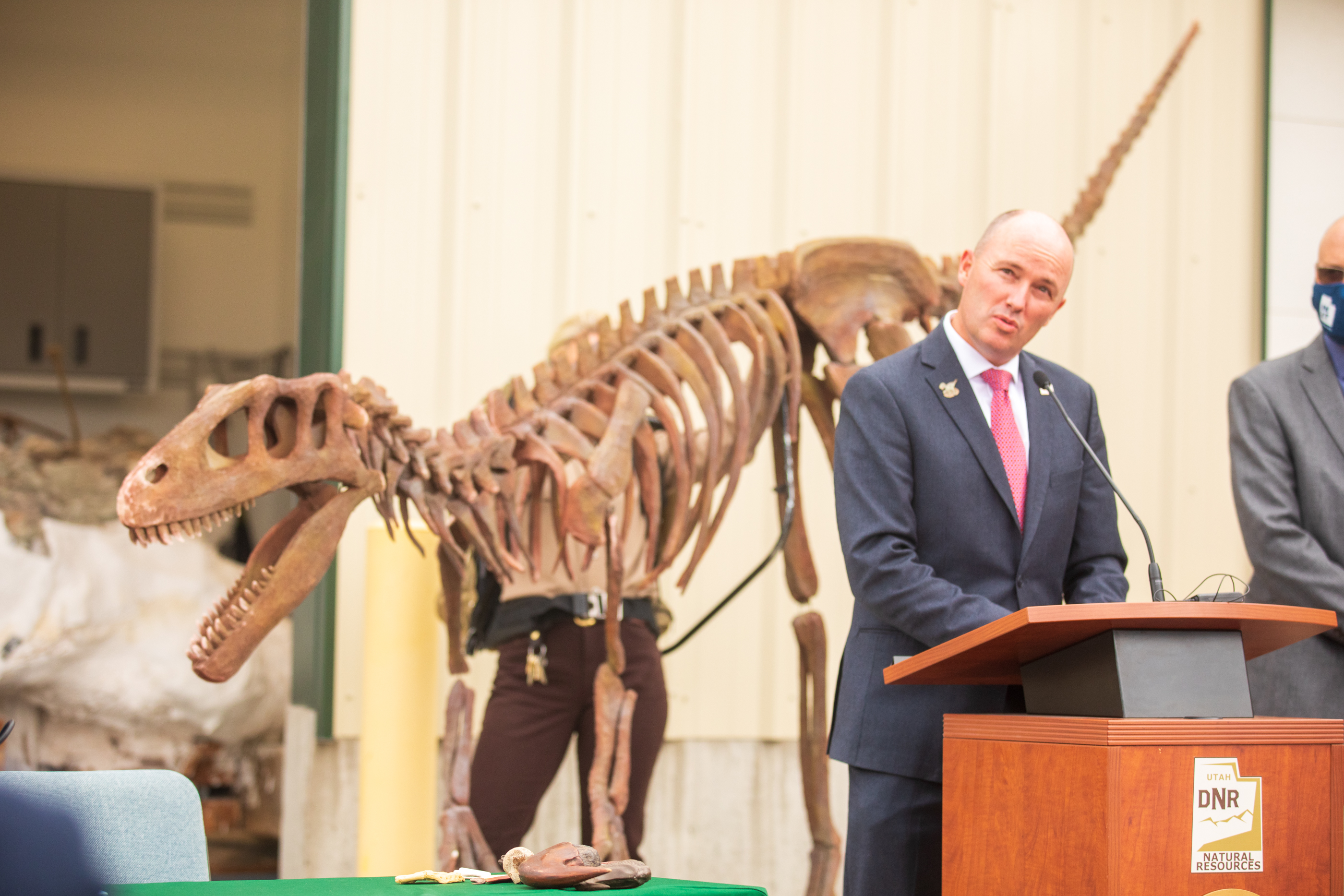 Gov. Spencer Cox speaks during a ceremony at the Utah Department of Natural Resources facility in Salt Lake City on Tuesday, April 13, 2021. A life-size replica of a Utahraptor is held up behind him.