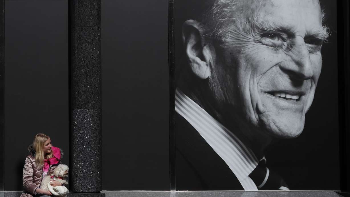 Isabella Disley and her dog Coco wait next to a portrait of Prince Philip, which adorns a window of a department store in Windsor, England, Friday, April 16, 2021. Prince Philip husband of Britain's Queen Elizabeth II died April 9, aged 99, his funeral will take place Saturday at Windsor Castle in St George's Chapel. (AP Photo/Alastair Grant)