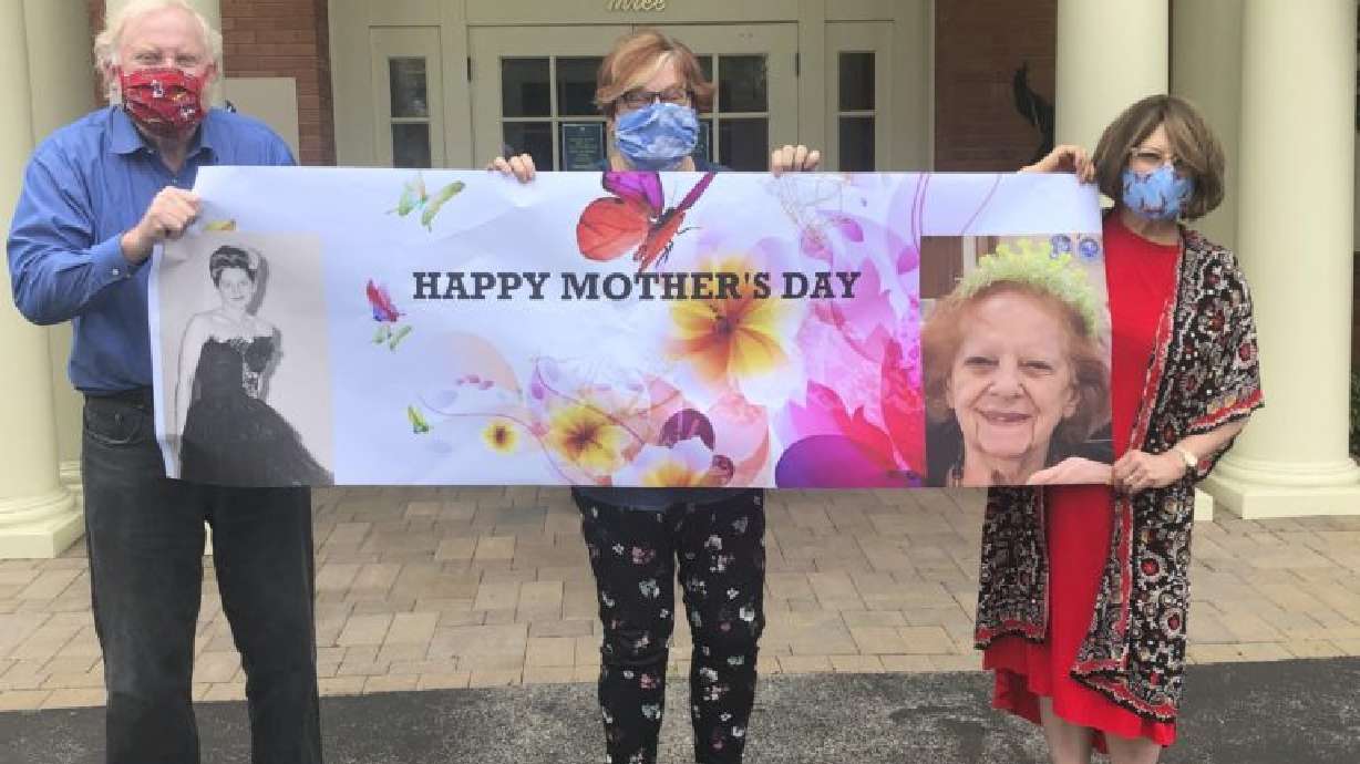 This May 3, 2020, photo released by Shelly Solomon shows, from left, Steve Turner and his sisters, Carla Paull and Lisa Fishman, holding up a Mother’s Day banner emblazoned with images of their mom, Beverly Turner, in front of her assisted living facility in Ladue, Missouri. In 2021, Utah officials say people who are fully vaccinated can celebrate Mother's Day in person without masks, even in long-term care facilities.