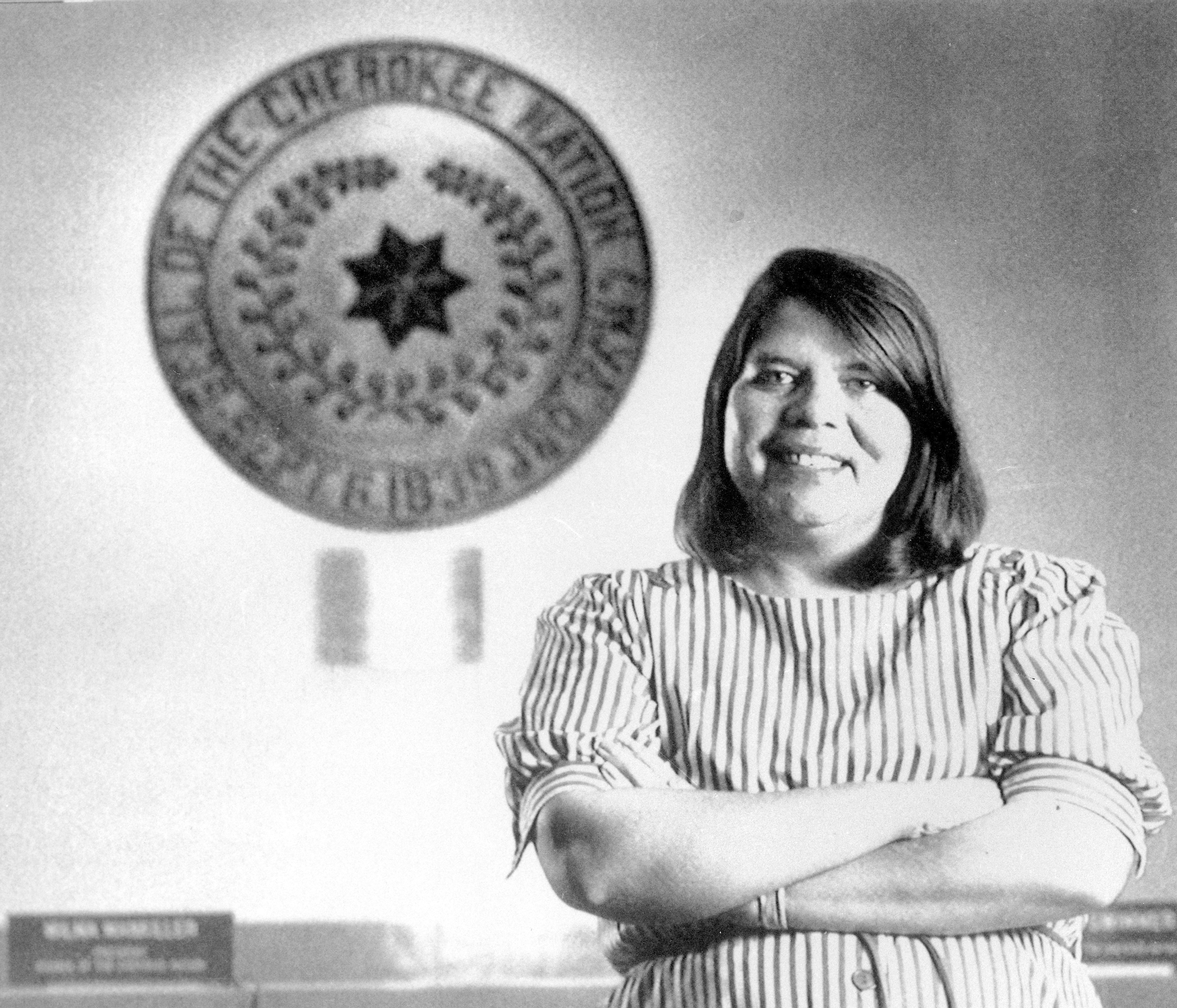 In this July 19, 1985, photo, Wilma Mankiller, the first woman elected chief of the Cherokee Nation, poses in front of the tribal emblem at the Cherokee Nation in Oklahoma. The U.S. Mint says Mankiller will be among the first five women honored in the new American Women Quarters Program, which starts in 2022 and continues until 2025. 
