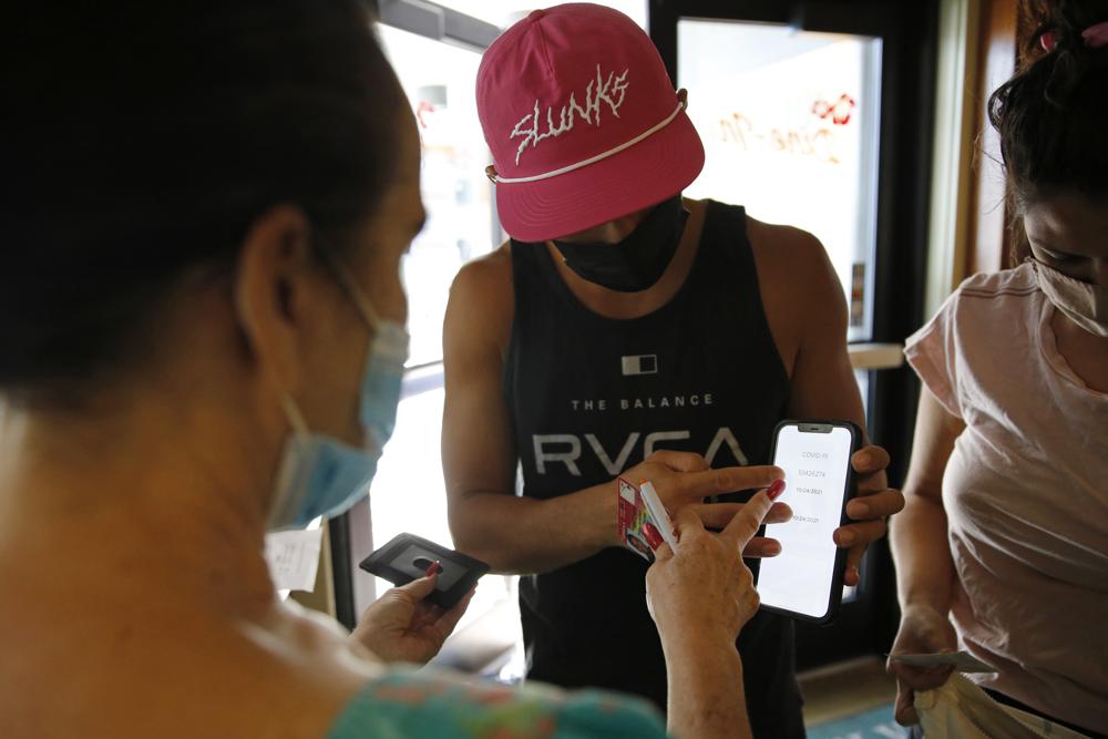 Highway Inn restaurant hostess Ku'uipo Lorenzo, left, checks a COVID-19 test result from Martin Day, center, as his wife, Ashley Day, right, prepares to show her vaccination card on Monday, in Honolulu. To comply with local mandates, the restaurant requires all indoor diners to show proof of vaccination or have a recent negative test result before being seated.
