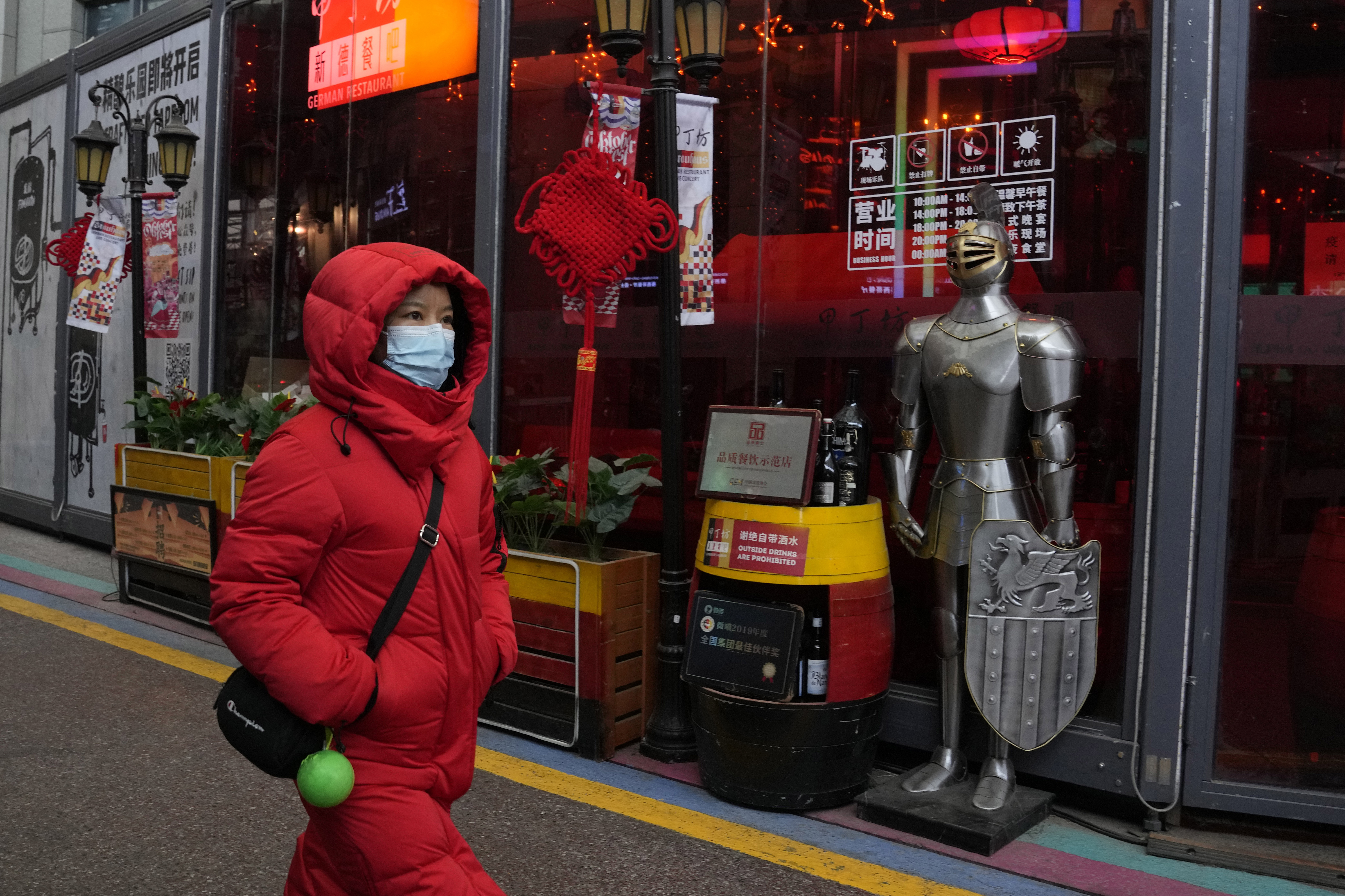 A woman wearing a mask to protect from the coronavirus walks past a coat of armour displayed outside a restaurant in Beijing, China, Friday, Jan. 21, 2022. The sweeping "zero-tolerance" policies that China has employed to protect its people and economy from COVID-19 may, paradoxically, make it harder for the country to exit the pandemic.