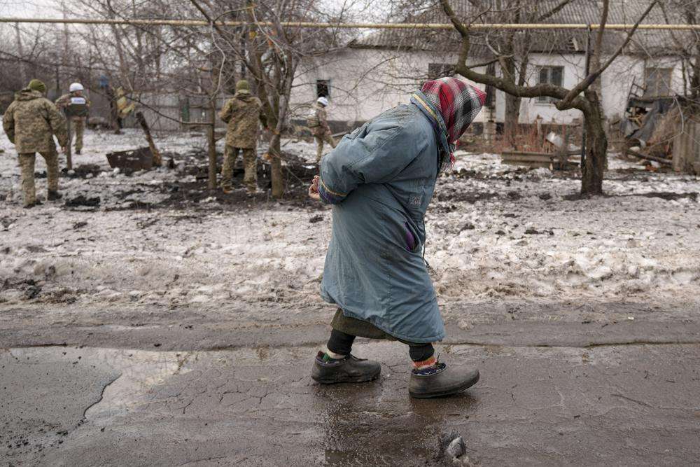 An elderly lady walks by as members of the Joint Centre for Control and Coordination on ceasefire of the demarcation line, survey a crater and damage to a house from artillery shell that landed in Vrubivka, one of the at least eight that hit the village today, according to local officials, in the Luhansk region, eastern Ukraine, Thursday. U.S. President Joe Biden warned that Russia could still invade Ukraine within days and Russia expelled the No. 2 diplomat at the U.S. Embassy in Moscow, as tensions flared anew in the worst East-West standoff in decades.