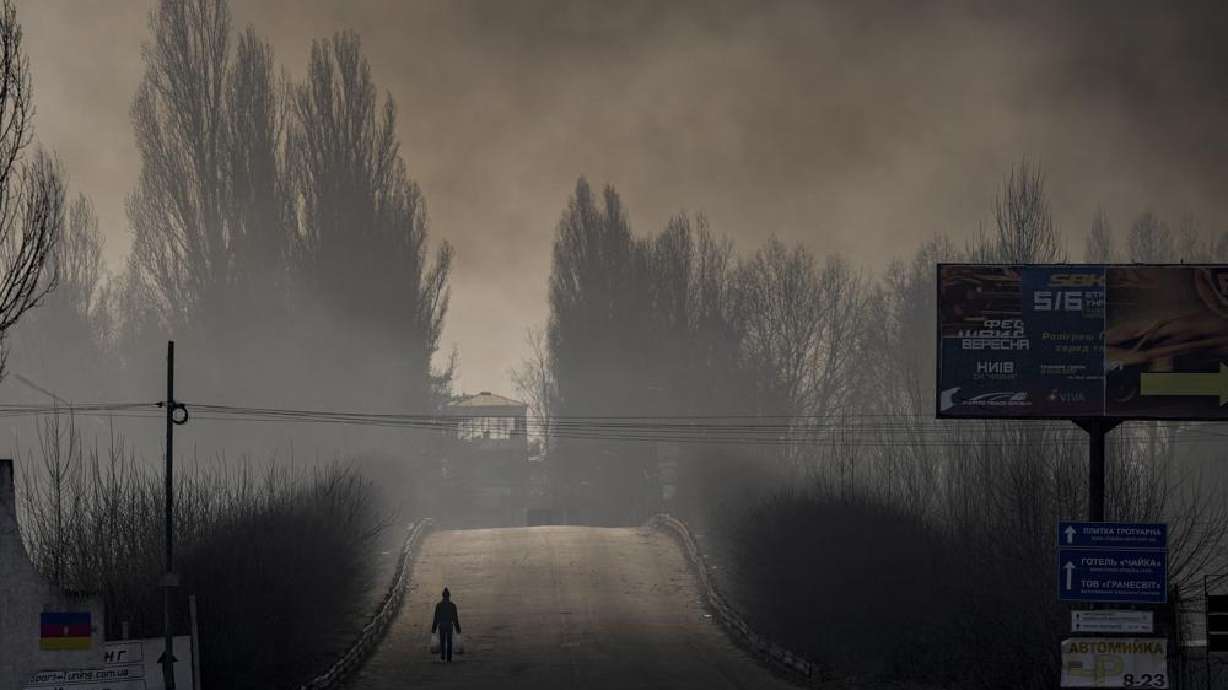 A man carries shopping bags as heavy smoke from a warehouse destroyed by Russian bombardment casts shadows on the road outside Kyiv, Ukraine, Thursday. Ukraine accused Moscow on Thursday of forcibly taking thousands of civilians from the shattered port city of Mariupol to Russia so that they can be used as “hostages” to pressure Kyiv to give up.