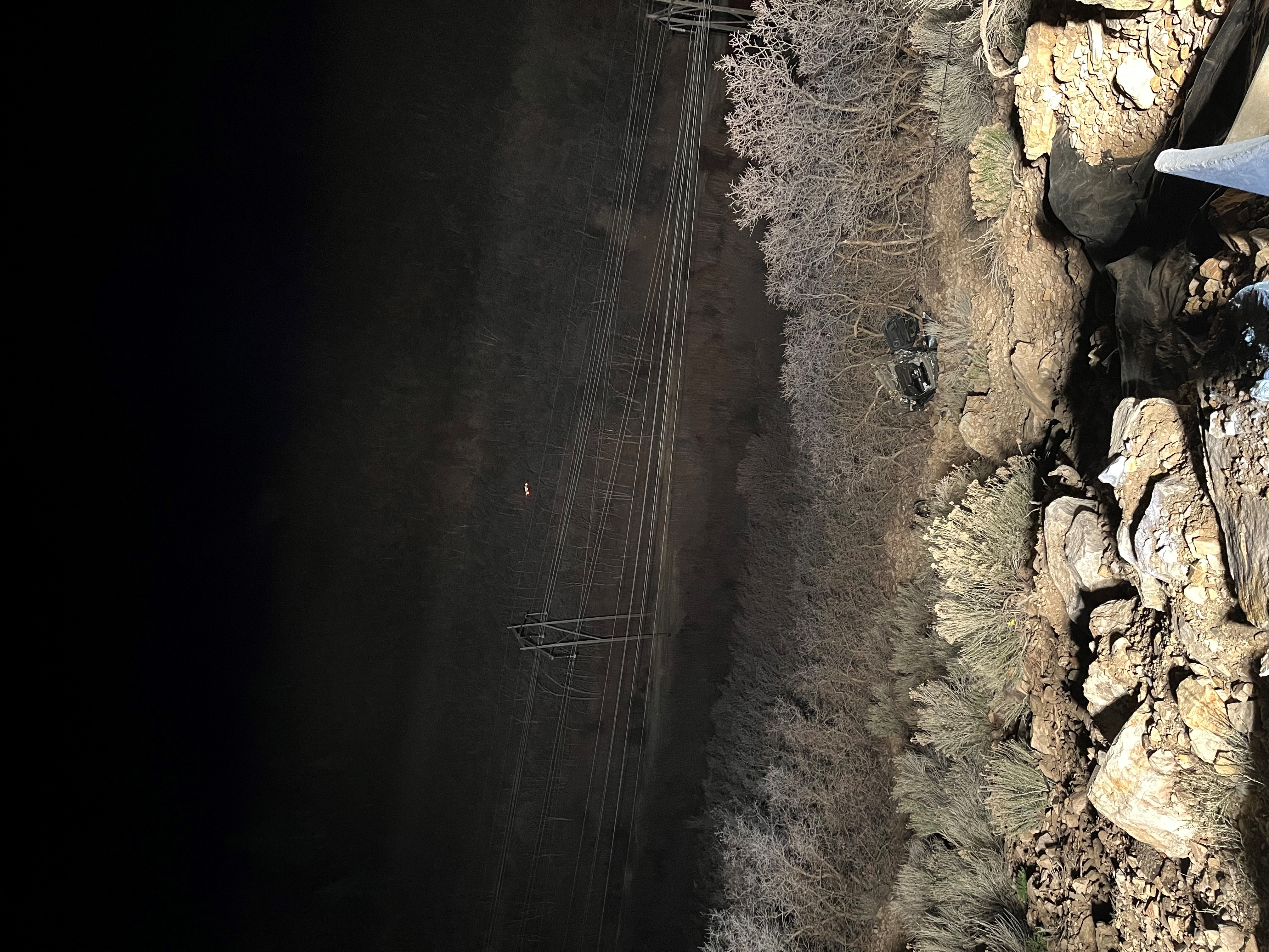 A mangled, dark-colored vehicle is shown stuck in a clump of trees to the right of some utility lines in North Ogden Canyon. The photo is taken from over the edge of the cliff the vehicle drove off of Thursday night. The Weber County Sheriff's Office said a woman was critically injured and a man also suffered injuries in the crash.