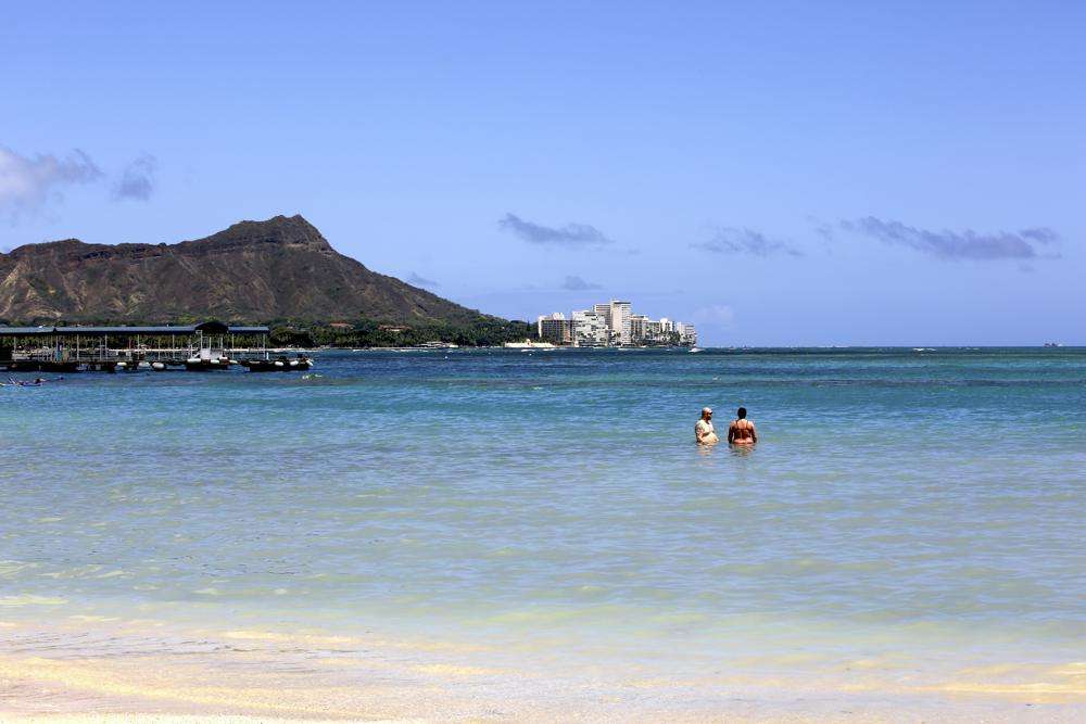Two people stand in the water off Waikiki Beach in Honolulu, Monday. A COVID surge is under way that is starting to cause disruptions for the year and Americans prepare for summer vacations. But the beaches beckoned and visitors have flocked to Hawaii, especially in recent months.