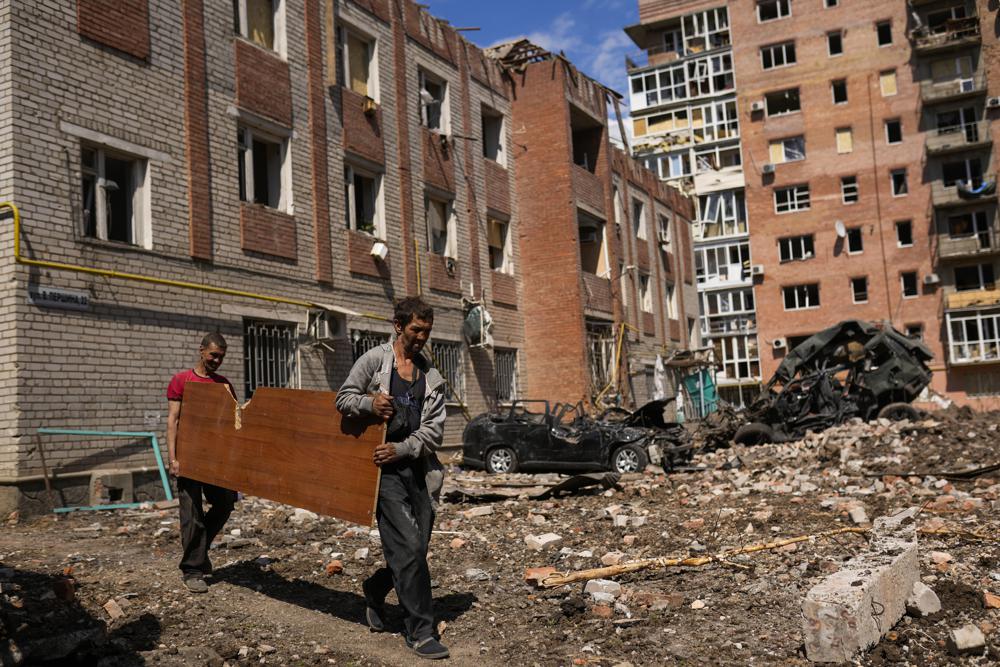 Two men carry a wooded panel next to heavily damaged buildings and destroyed cars in a Russian bombing in Bakhmut, eastern Ukraine, Tuesday. Curfews, checkpoints and fortifications have become commonplace after 90 days of war in Ukraine.