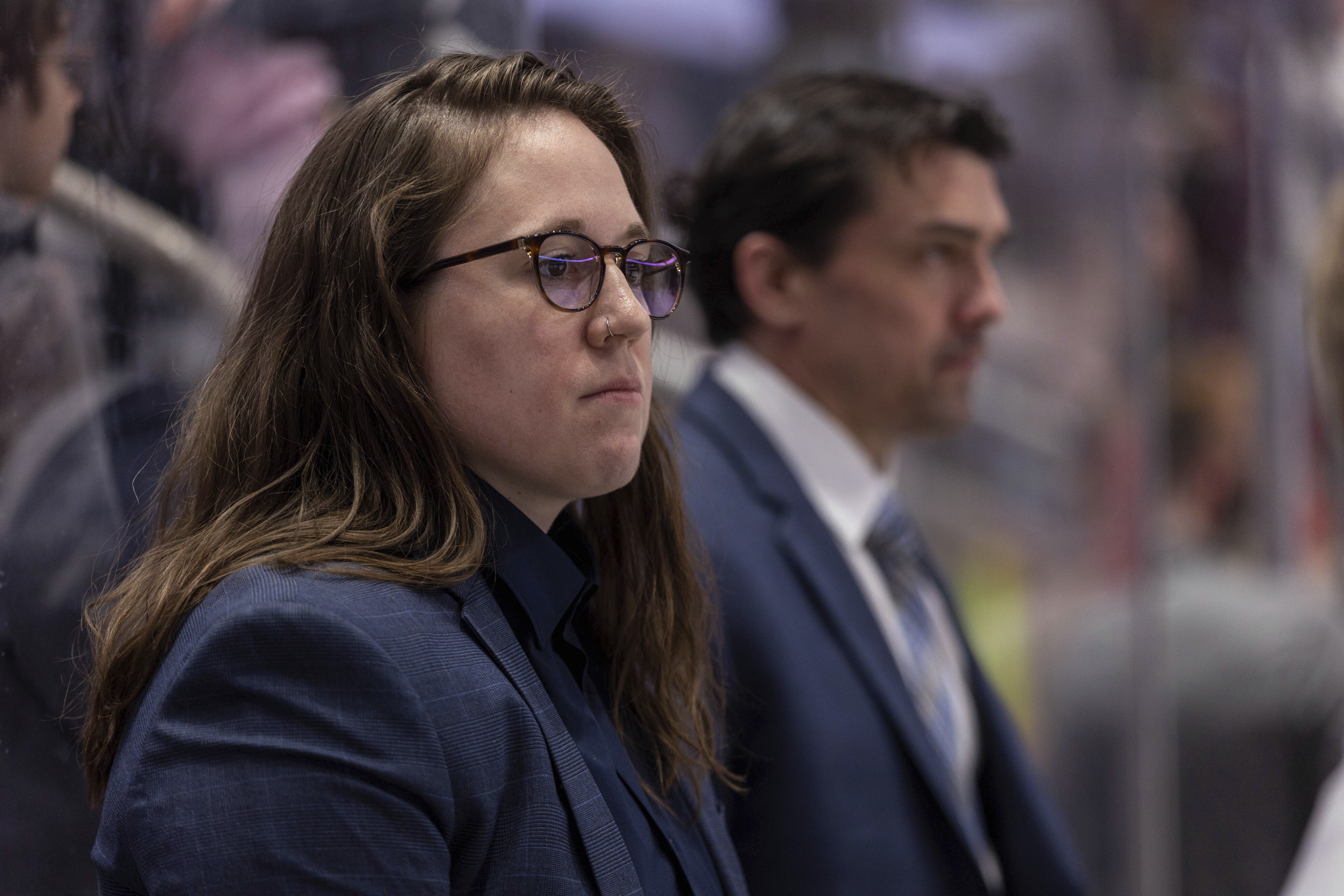 Emily Engel-Natzke stands on the bench in front of associate goaltending coach Alex Westlund during pregame warmups before the Hershey Bears played the Springfield Thunderbirds in an American Hockey League game on Sunday, April 10, 2022, in Hershey, Pa. Engel-Natzke was promoted from Hershey to video coach of the Washington Capitals on Thursday, June 30, 2022, making her the first woman to hold that position in the National Hockey League. 