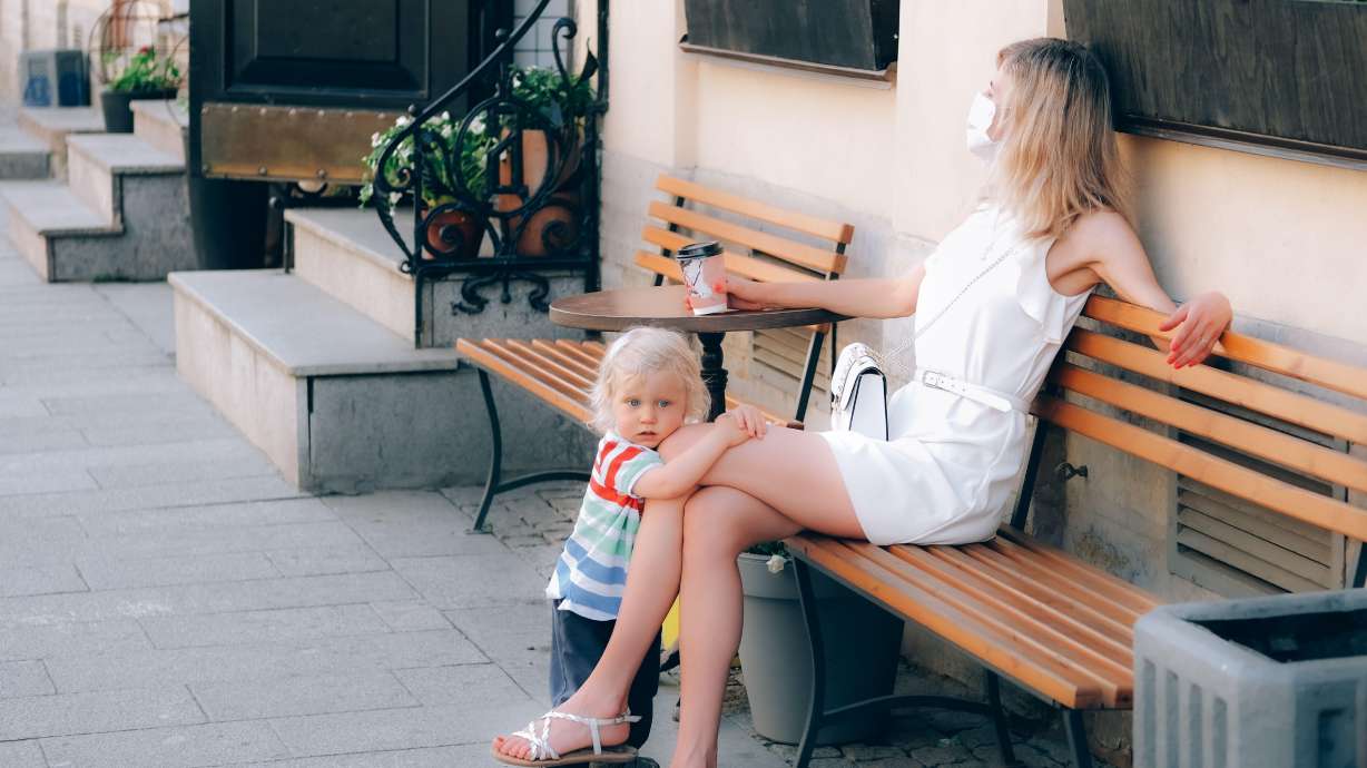 A young woman wearing a medical mask sitting on the bench of outdoor cafe with her child. Resilience is tough when it feels like your difficulties will never end.