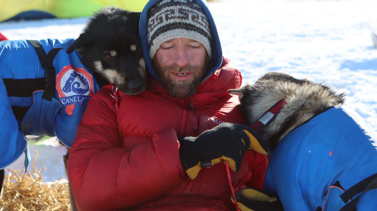 FILE - Musher Jessie Holmes takes a break from cooking his dogs a meal to nuzzle with two wheel dogs at the Ophir checkpoint during the Iditarod Trail Sled Dog Race on March 10, 2021, in Alaska. The television star and Iditarod musher sustained injuries this week when helping clean up storm damage along the Bering Sea coast. Jessie Holmes, who since 2015 has starred in "Life Below Zero," about life in rural Alaska produced by National Geographic TV, was injured by falling debris in a building in the community of Golovin on Wednesday, Sept. 28, 2022, the Anchorage Daily News reported. He was flown for treatment to Nome and then sent on to an Anchorage hospital.