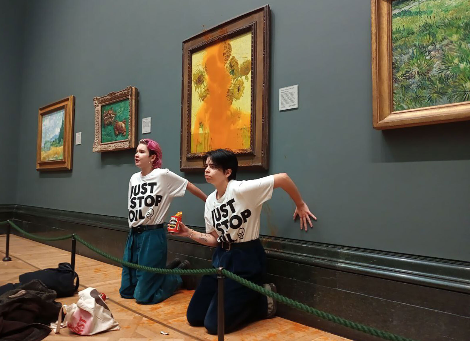 In this image released by the Just Stop Oil organization, two women are seen standing in front of Van Gogh's "Sunflowers" (1888) in a London gallery Friday. The anti-fossil fuel protesters were filmed throwing tomato soup on the artwork Friday and have since been charged with criminal damage offenses.