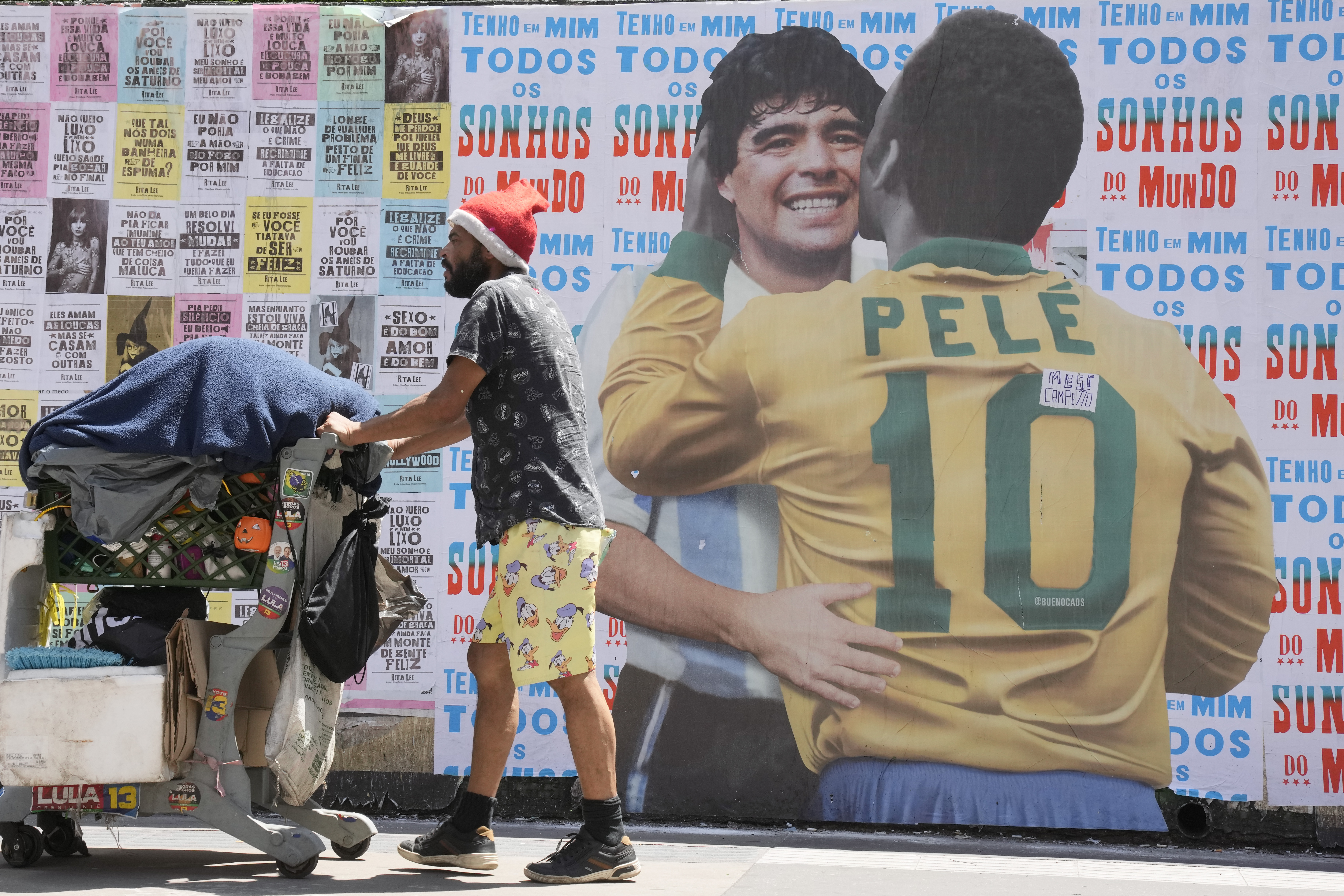 A man pushes a cart loaded with garbage for recycling in front of a mural depicting Brazilian soccer legend Pele embracing late Argentinean soccer star Diego Maradona in Sao Paulo, Brazil, Saturday, Dec. 24, 2022. Pele is hospitalized to continue his cancer treatment.