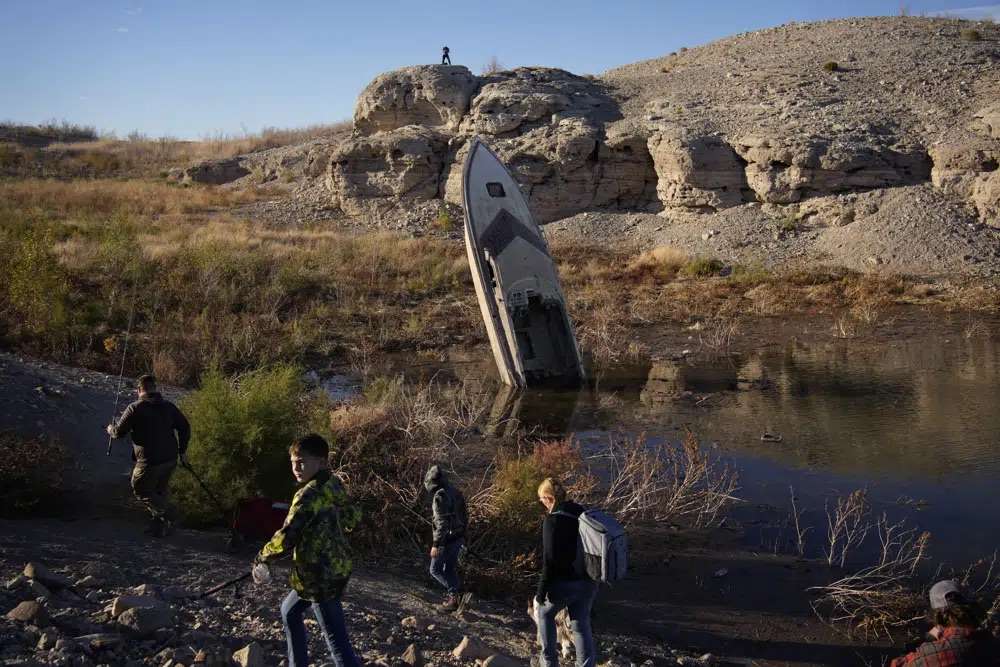 People walk by a formerly sunken boat standing upright into the air with its stern buried in the mud along the shoreline of Lake Mead at the Lake Mead National Recreation Area, Friday near Boulder City, Nev. More than 10% of the water carried by the Colorado River evaporates, leaks or spills as the 1,450-mile powerhouse river of the West flows through the region’s dams, reservoirs and open-air canals.