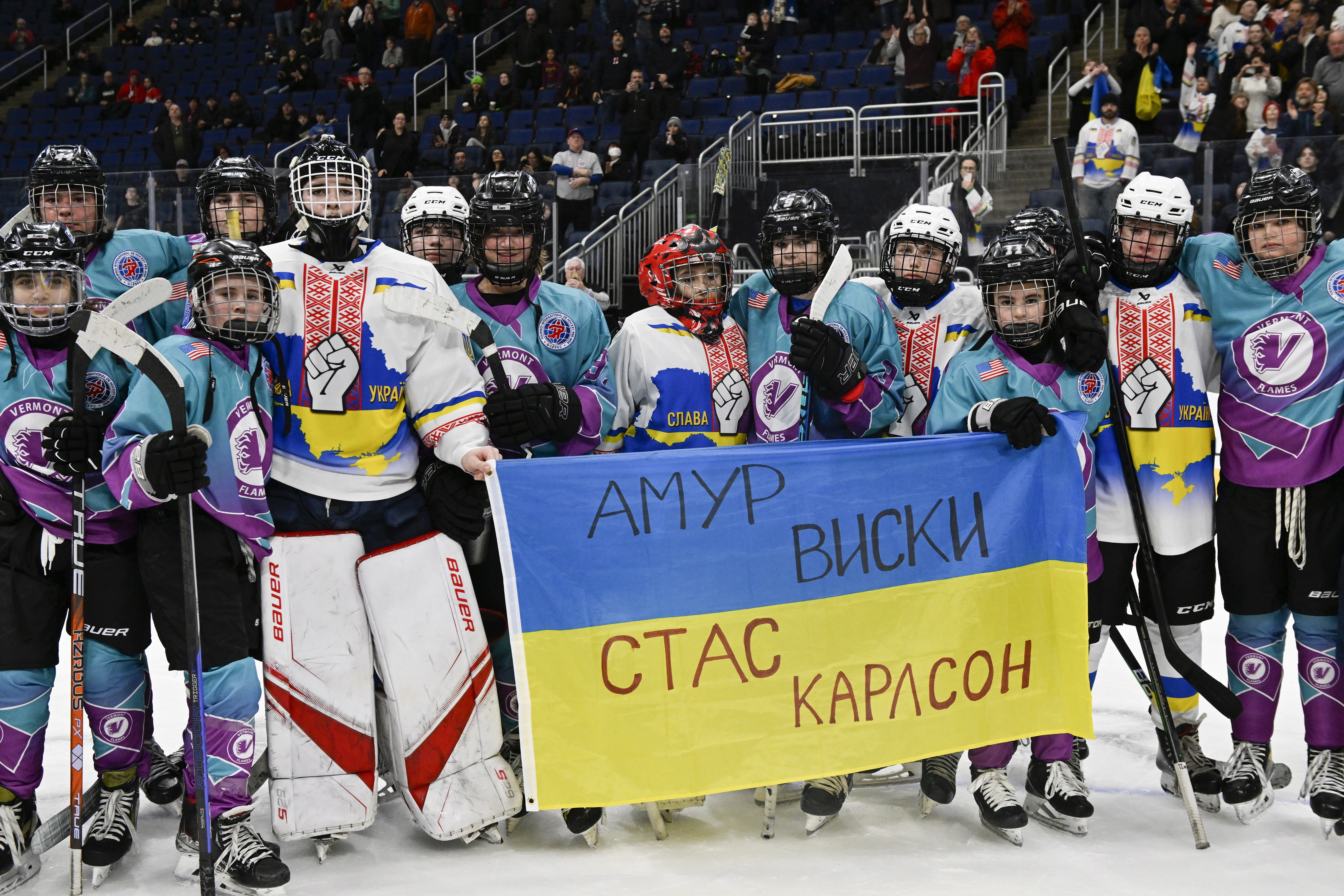 Vermont Flames Academy and Ukraine hockey players stand together after a game at the International Peewee Tournament in Quebec City, Friday, Feb. 17, 2023. Ukraine lost 2-1 to Vermont Flames Academy. Written on the top of the flag is the name of Amur Whiskey, who died in the war and was the father of one of the players on the team. The bottom of the flag has the name Stas Karlson, who is the father of another player and is still fighting.