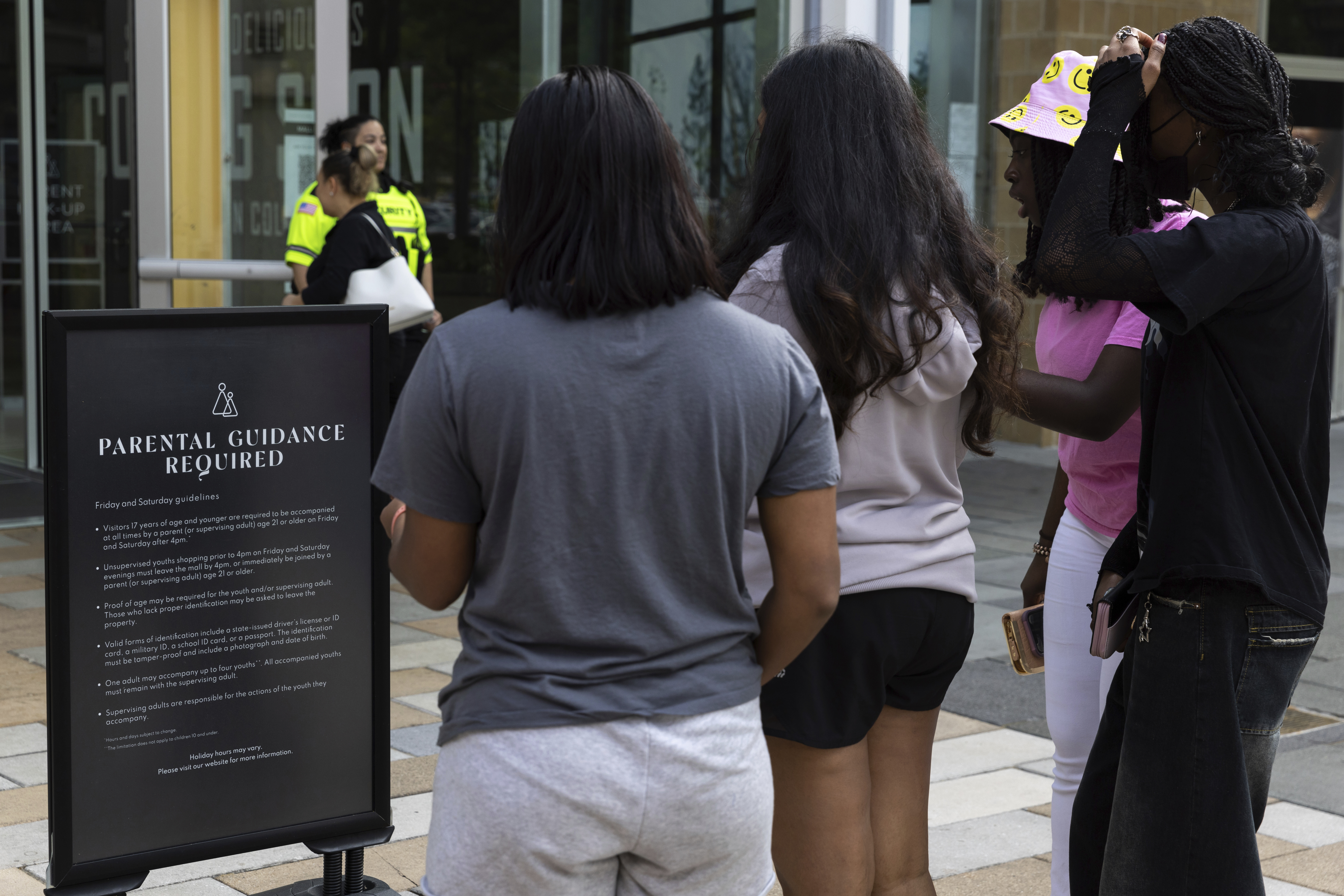 A security guard stands by as teens read a sign explaining the Mall in Columbia's "Parental Guidance Required" program May 12, 2023, in Columbia, Md. The program requires that all visitors under 18 be accompanied by an adult who is at least 21-years-old after 4 p.m. on Fridays and Saturdays.