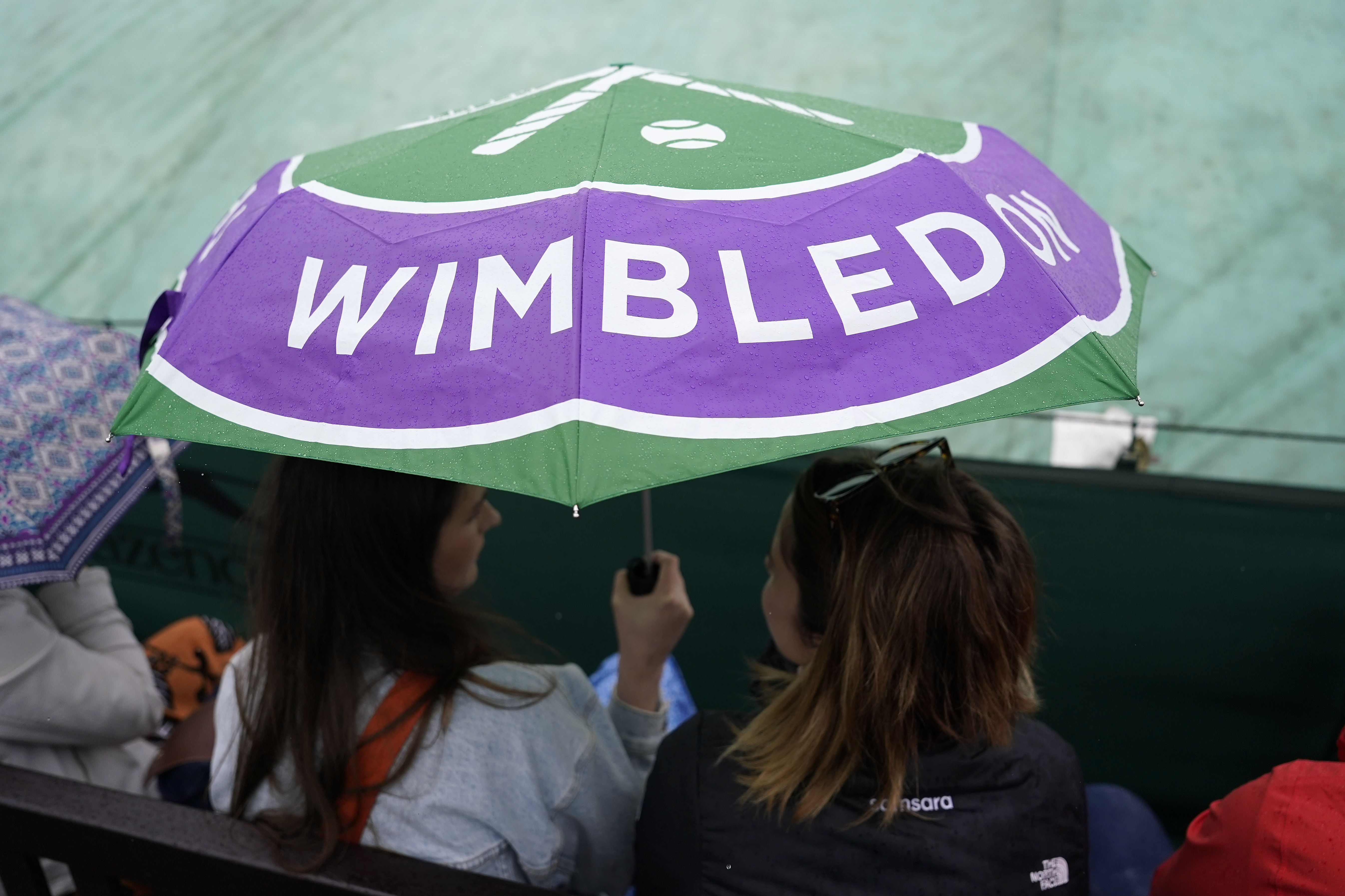 Spectators sit under an umbrella on a covered court after rain delayed the start of play on day three of the Wimbledon tennis championships in London, Wednesday, July 5, 2023.