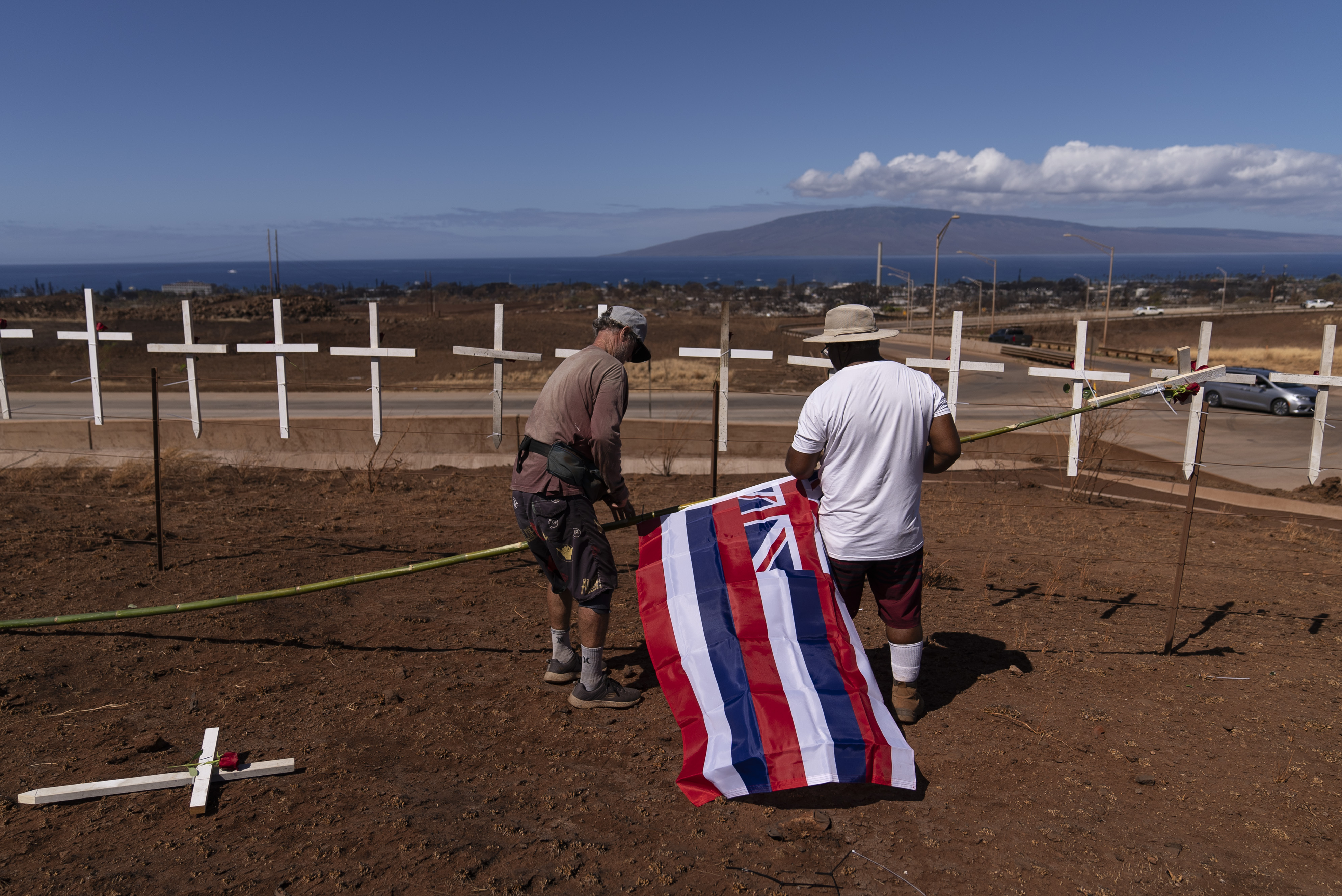 Matt Schweitzer and Soakai Taufa hang a Hawaiian flag honoring the victims killed in Lahaina, Hawaii, wildfires. Two weeks after the deadly wildfire, between 500 and 1,000 people remain unaccounted for. 