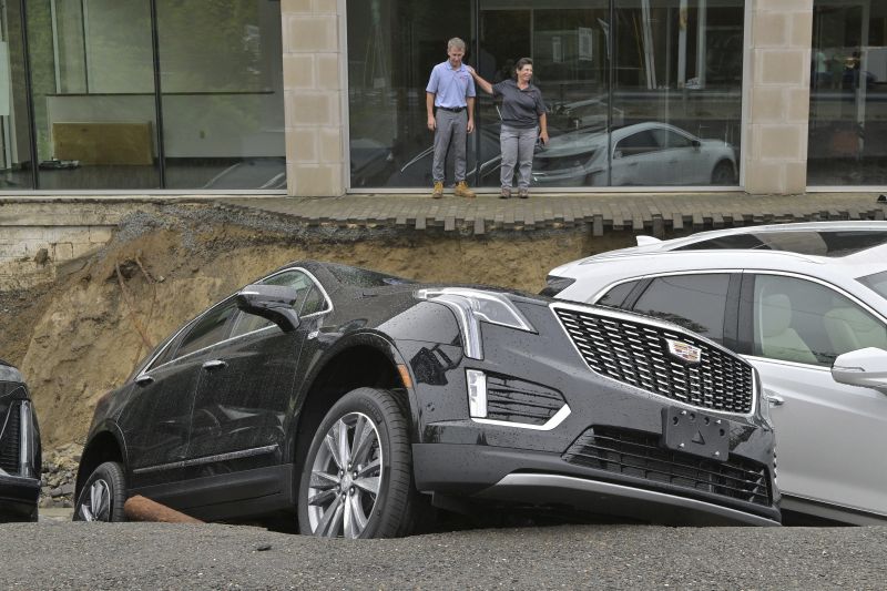 From left, car dealership owner Rick Durand Owner of Durand Cadillac and controller Michelle Bettez react beside three vehicles that fell into a sinkhole that was washed out of his car dealership Tuesday, in Leominster, Mass., after more than nine inches of rain fell overnight.