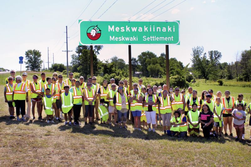 Members of the Meskwaki Nation along with employees from the Iowa Department of Transportation and Federal Highway Administration stand in front of a sign on U.S. Highway 30 that features the Meskwaki Nation's own spelling of the tribe, Meskwakiinaki on July 13, 2022.