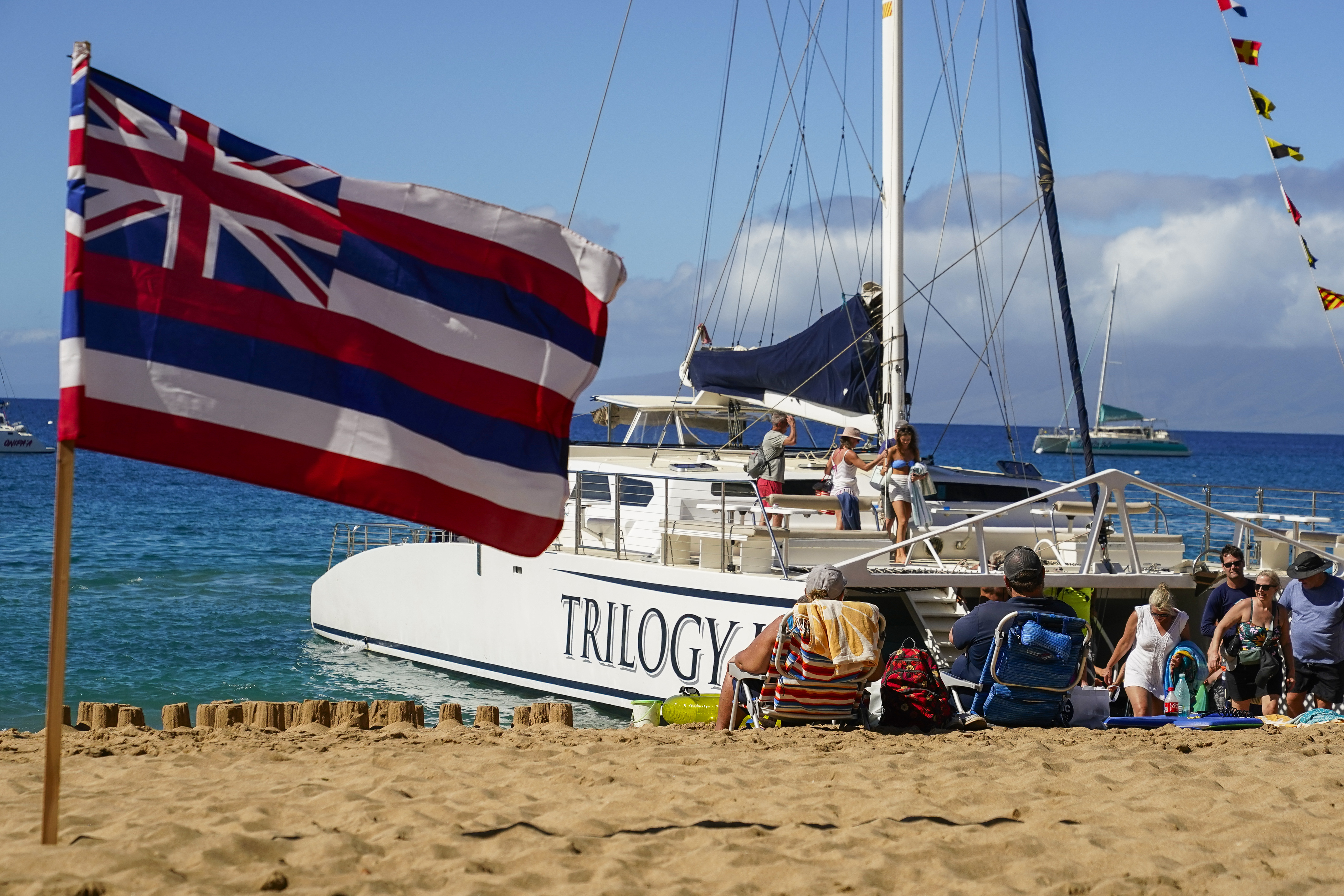 Tourists get off of a Trilogy Excursions boat arriving on Kaanapali Beach in front of a flag of Hawaii planted in the sand Dec. 6,  in Lahaina, Hawaii. Residents and survivors are still dealing with the aftermath of the August wildfires in Lahaina.