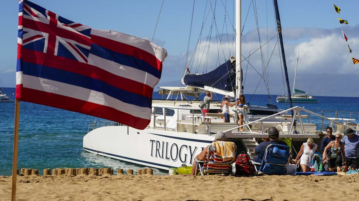 Tourists get off of a Trilogy Excursions boat arriving on Kaanapali Beach in front of a flag of Hawaii planted in the sand Dec. 6, in Lahaina, Hawaii. Residents and survivors are still dealing with the aftermath of the August wildfires in Lahaina.
