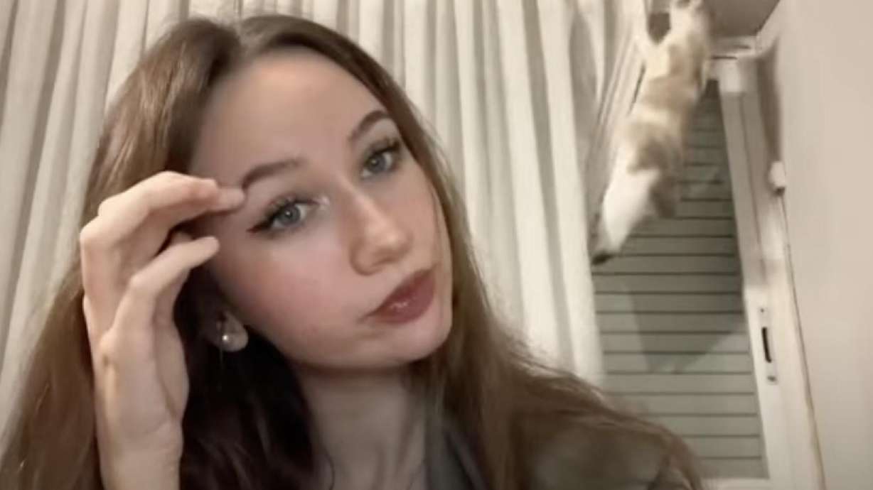 Woman pauses during filming in surprise at a cat climbing curtain behind her.