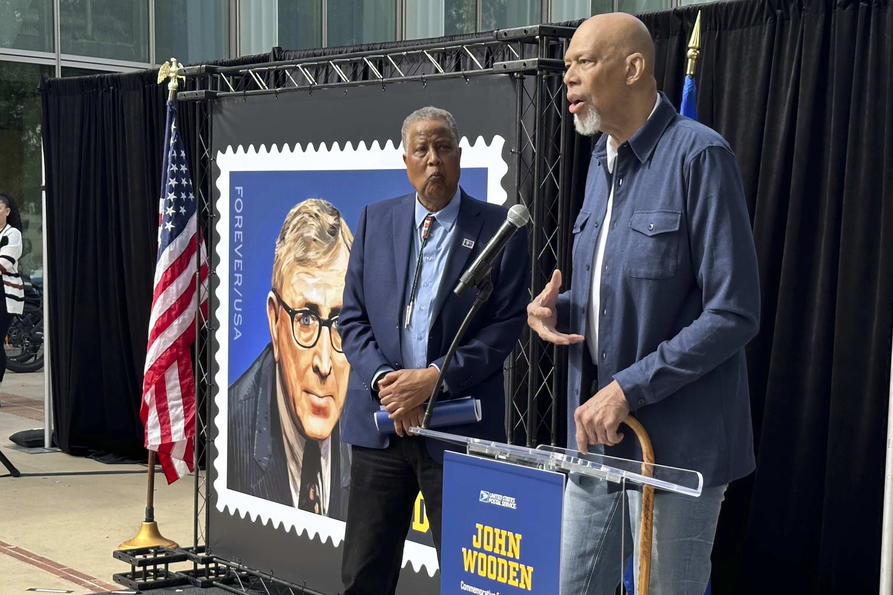 Jamaal Wilkes, left, listens as Kareen Abdul-Jabbar speaks at the first-day-of-issue ceremony for the John Wooden forever stamp on the UCLA campus in Los Angeles, Calif., on Saturday, Feb. 24, 2024. Wilkes and Abdul-Jabbar both played for Wooden, who coached UCLA to a record 10 national basketball championships. Wooden died in 2010 at age 99. 