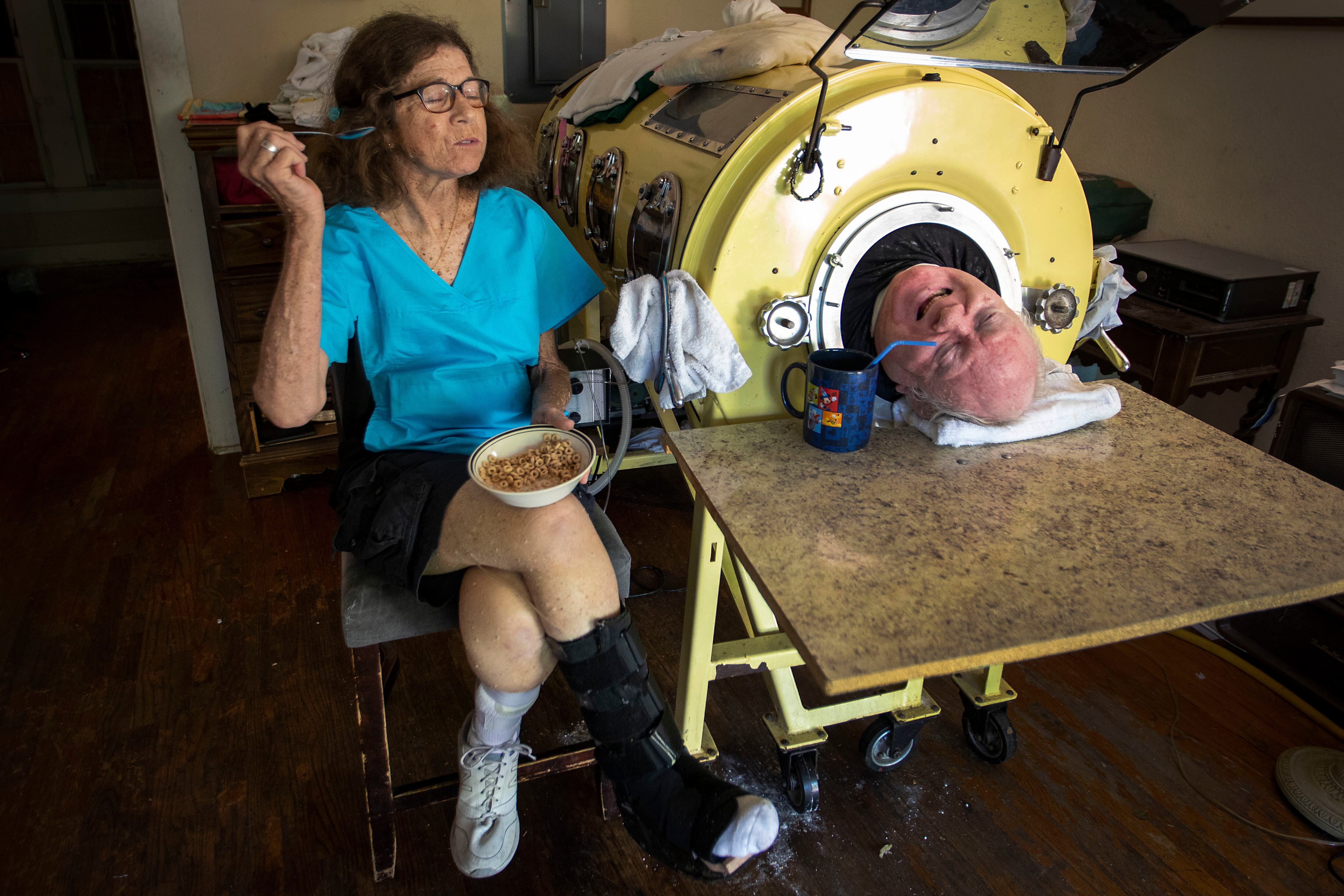 In this April 27, 2018, photo, attorney Paul Alexander chats with caregiver and friend Kathryn Gaines as he drinks coffee and she eats breakfast beside his iron lung at his home in Dallas. Alexander died Monday.