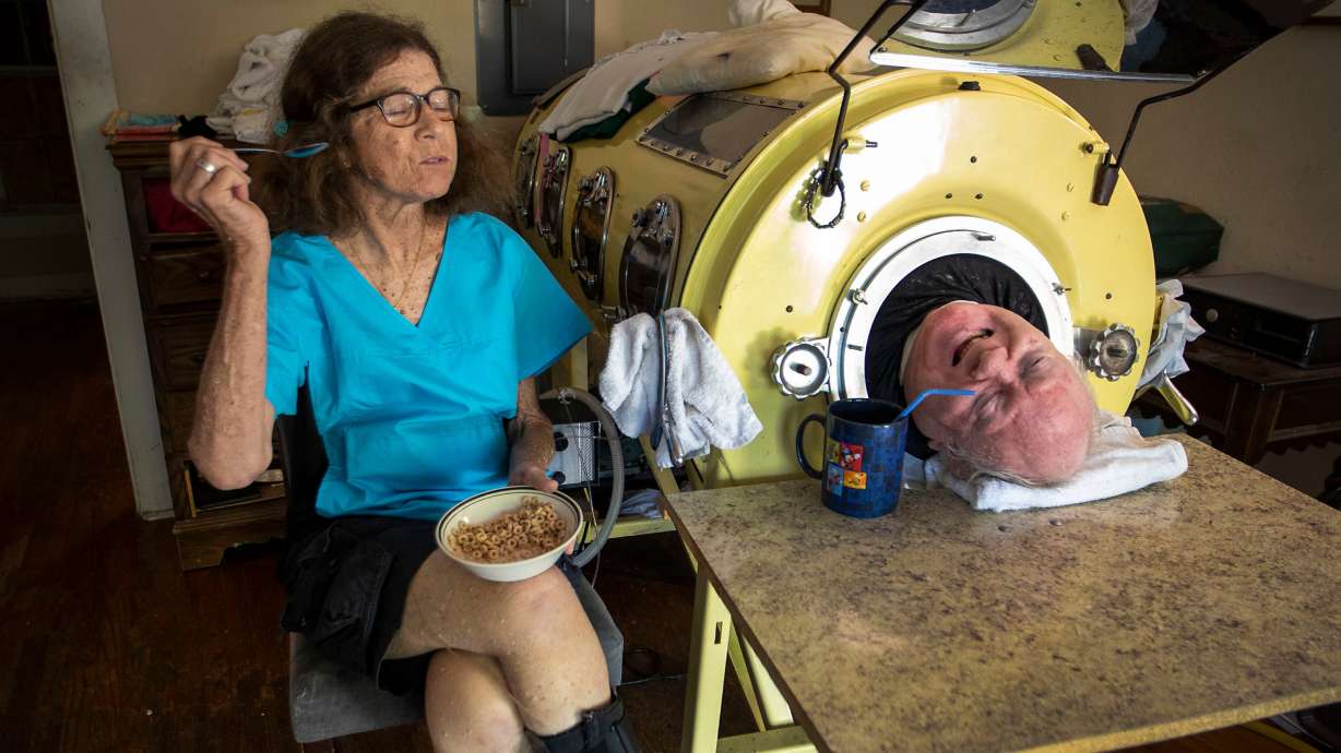 In this April 27, 2018, photo, attorney Paul Alexander chats with caregiver and friend Kathryn Gaines as he drinks coffee and she eats breakfast beside his iron lung at his home in Dallas. Alexander died Monday.