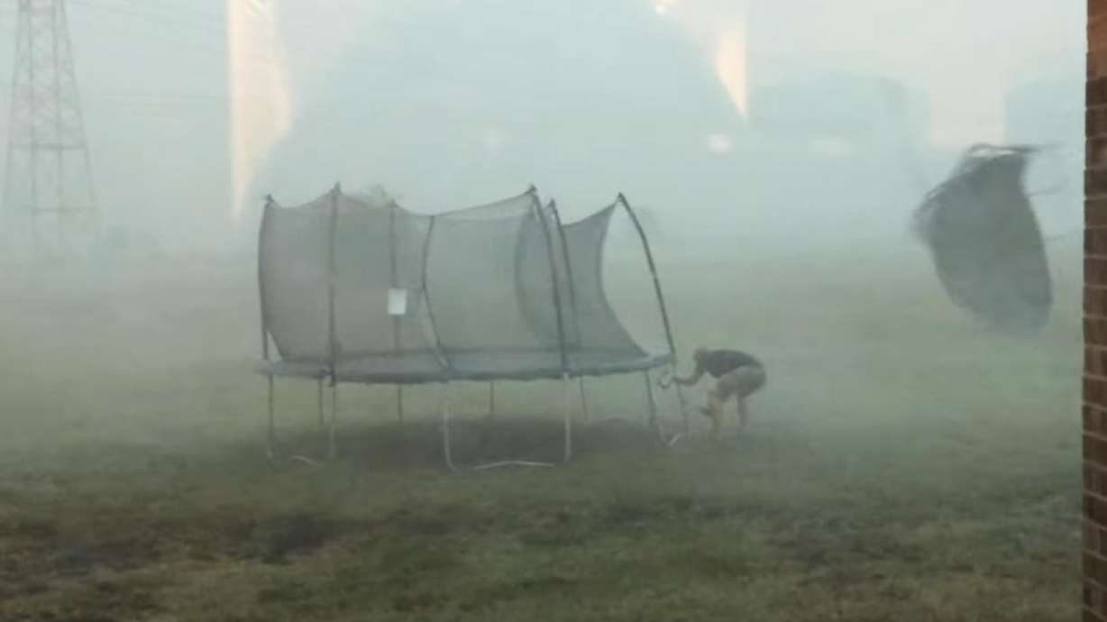 A man hammers stakes around a trampoline in a storm when another trampoline flies behind him in the wind. The rain picks up and the trampoline he's hammering down flies away and knocks him over.
