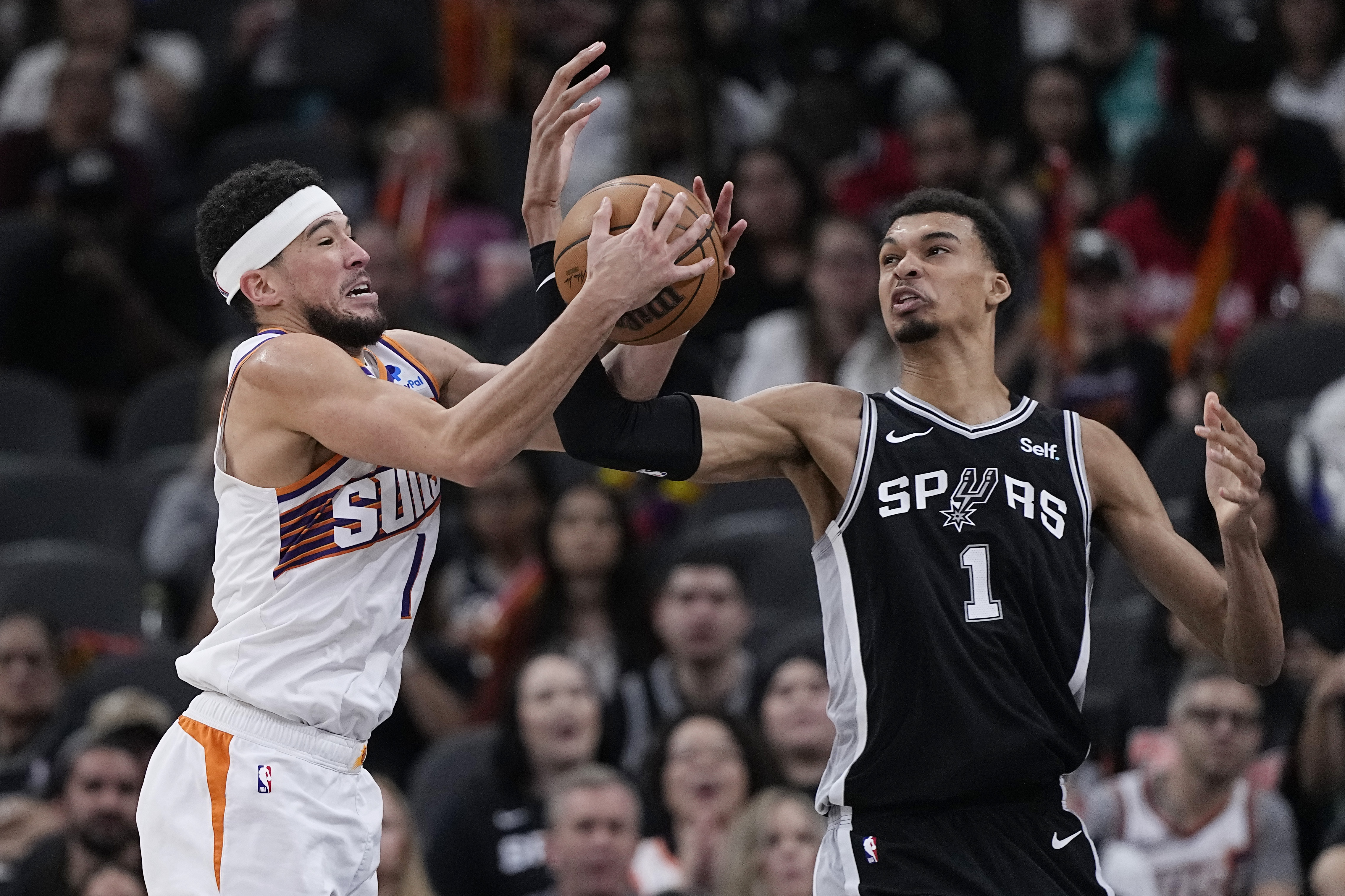 Phoenix Suns guard Devin Booker, left, and San Antonio Spurs center Victor Wembanyama, right, reach for a rebound during the second half of an NBA basketball game in San Antonio, Saturday, March 23, 2024.