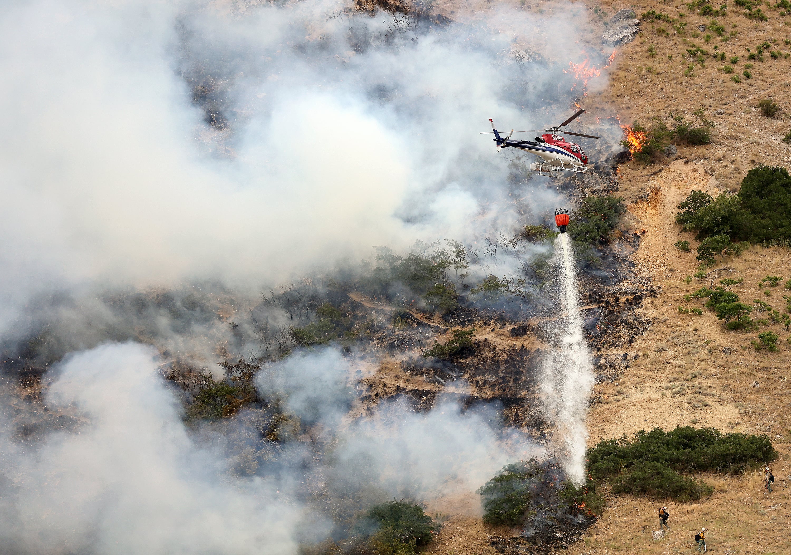 Firefighters battle a wildfire from the ground as a helicopter drops water above them in Springville on Aug. 1, 2022. The fire started when a man tried to burn a spider with a lighter.