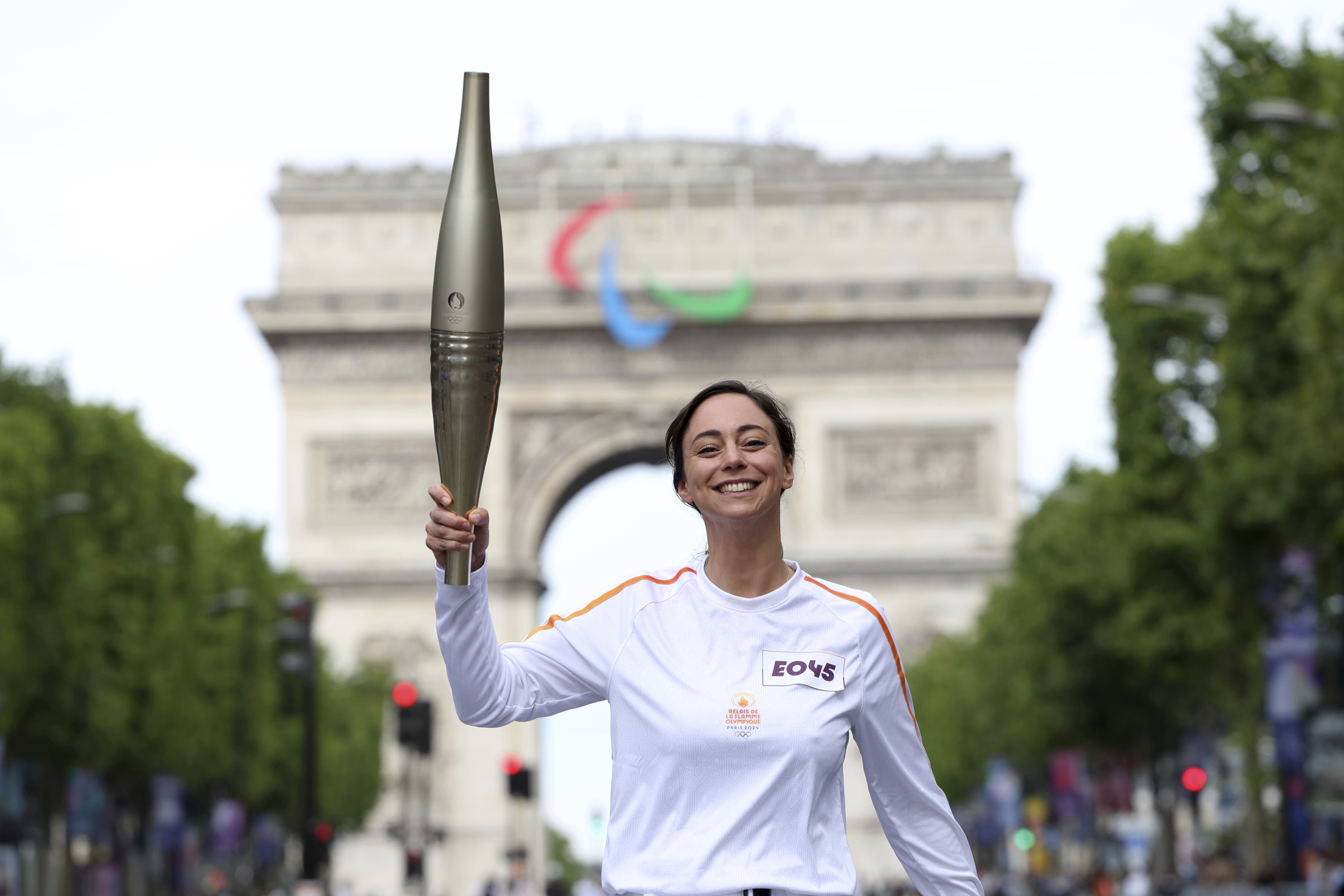 Torchbearer Nina Metayer, a French pastry maker, holds the torch on the Champs-Elysees avenue, with the Arc de Triomphe in background, Monday, July 15, 2024 in Paris, just 11 days before the French capital hosts exceptionally ambitious and high-security Paris Olympic Games. 